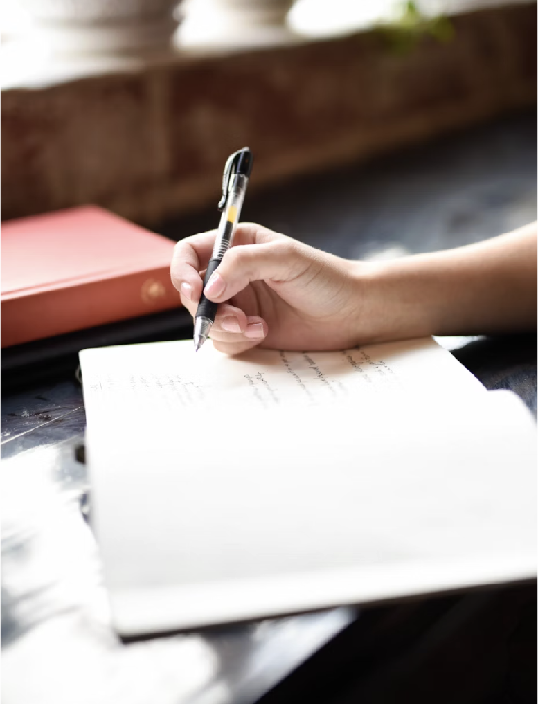 A person writing in a notebook with a black and yellow pen, on a dark table near a window with sunlight.