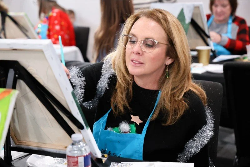 Woman painting on an easel at holiday-themed event with other people in the background.