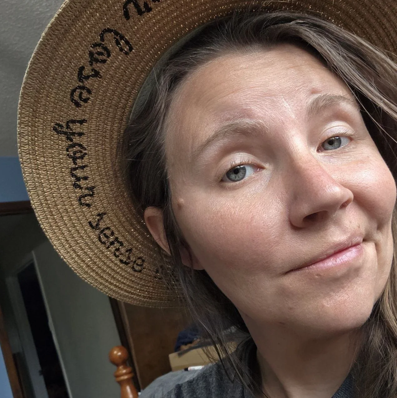 A woman with light skin and light blue eyes wearing a wide-brimmed straw hat with black writing, smiling softly indoors.