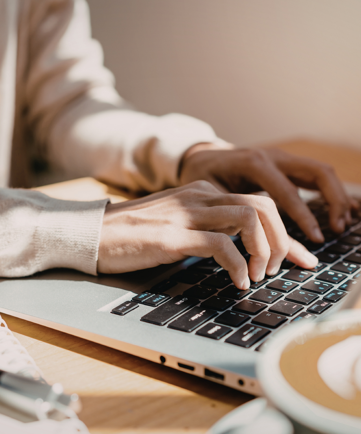 Two people typing on a laptop keyboard with one hand each, sitting at a wooden table with a white mug nearby.