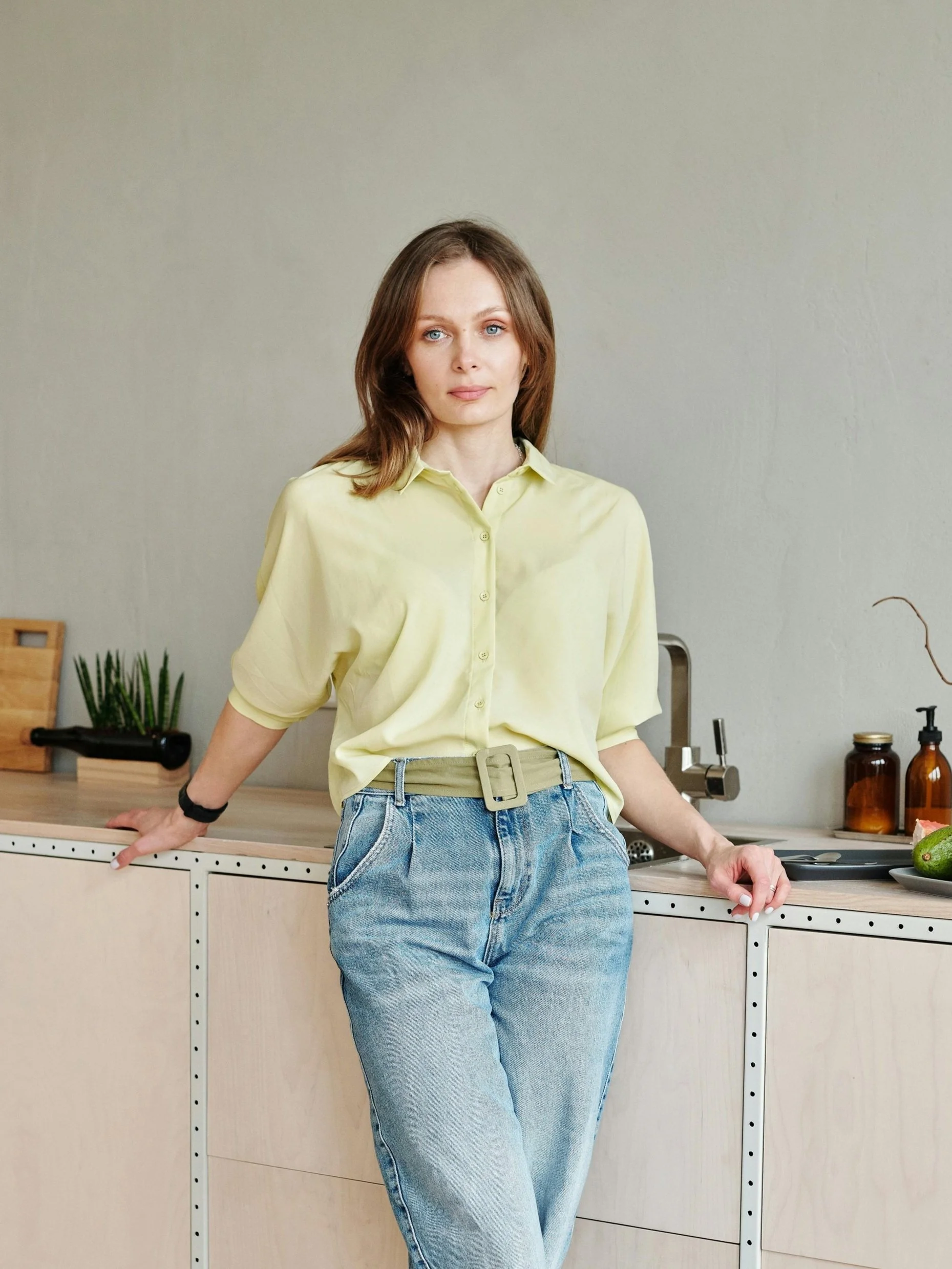A person wearing a pale yellow shirt and light blue jeans leaning against a kitchen countertop. The kitchen has a modern design with light wooden cabinetry, a plant, a cutting board, a jar, a soap dispenser, and a green avocado.