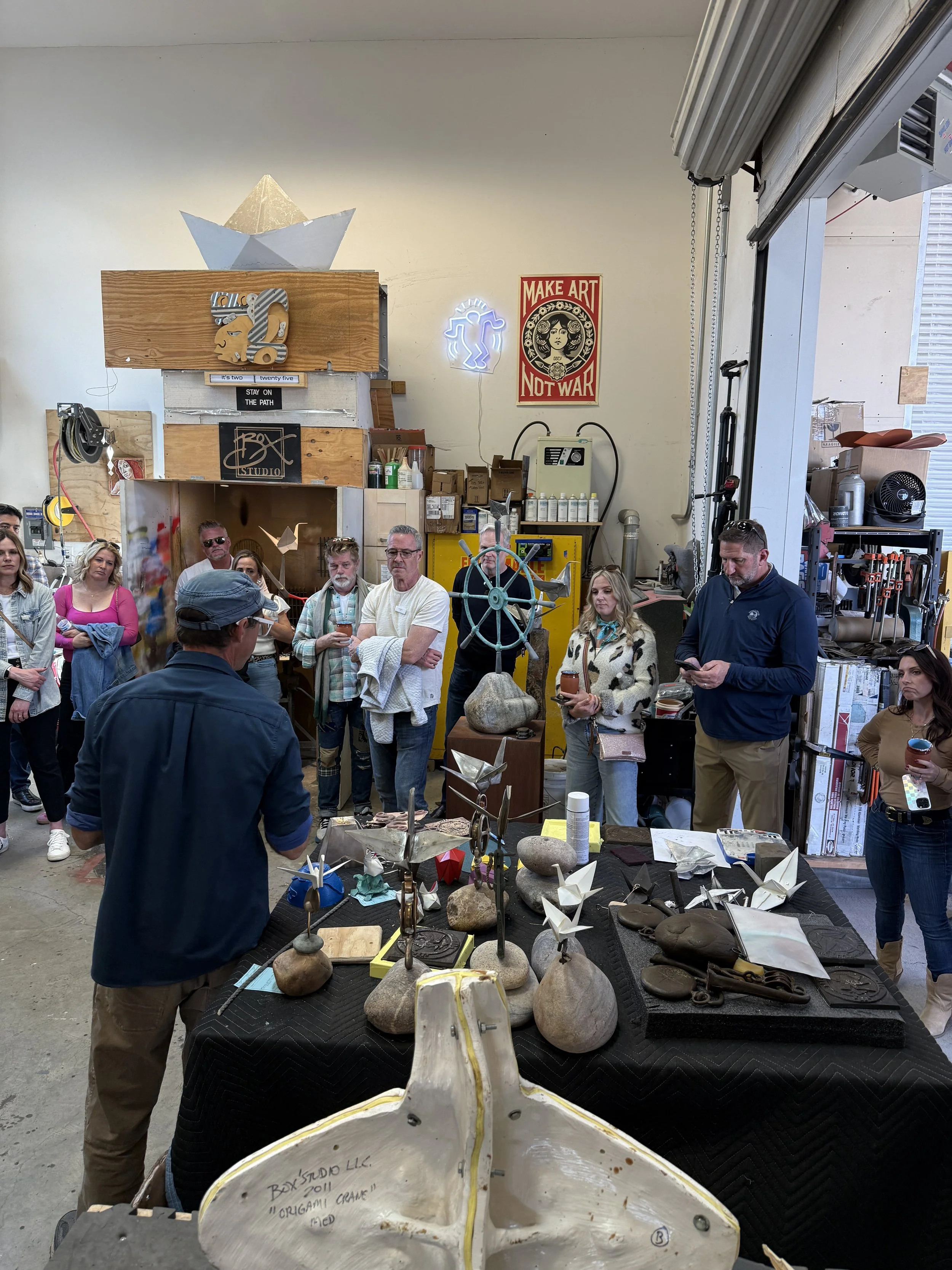 A group of people gathered around a table displaying various metal art sculptures and rocks, inside an industrial-style art studio.
