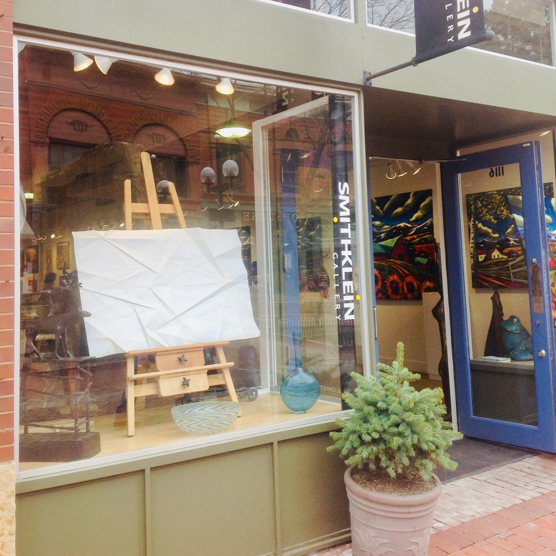 A storefront window with artwork displayed inside and a potted evergreen tree outside. The window has a green frame and the interior shows paintings, one resembling Van Gogh's starry night. The scene is outside on a sidewalk.