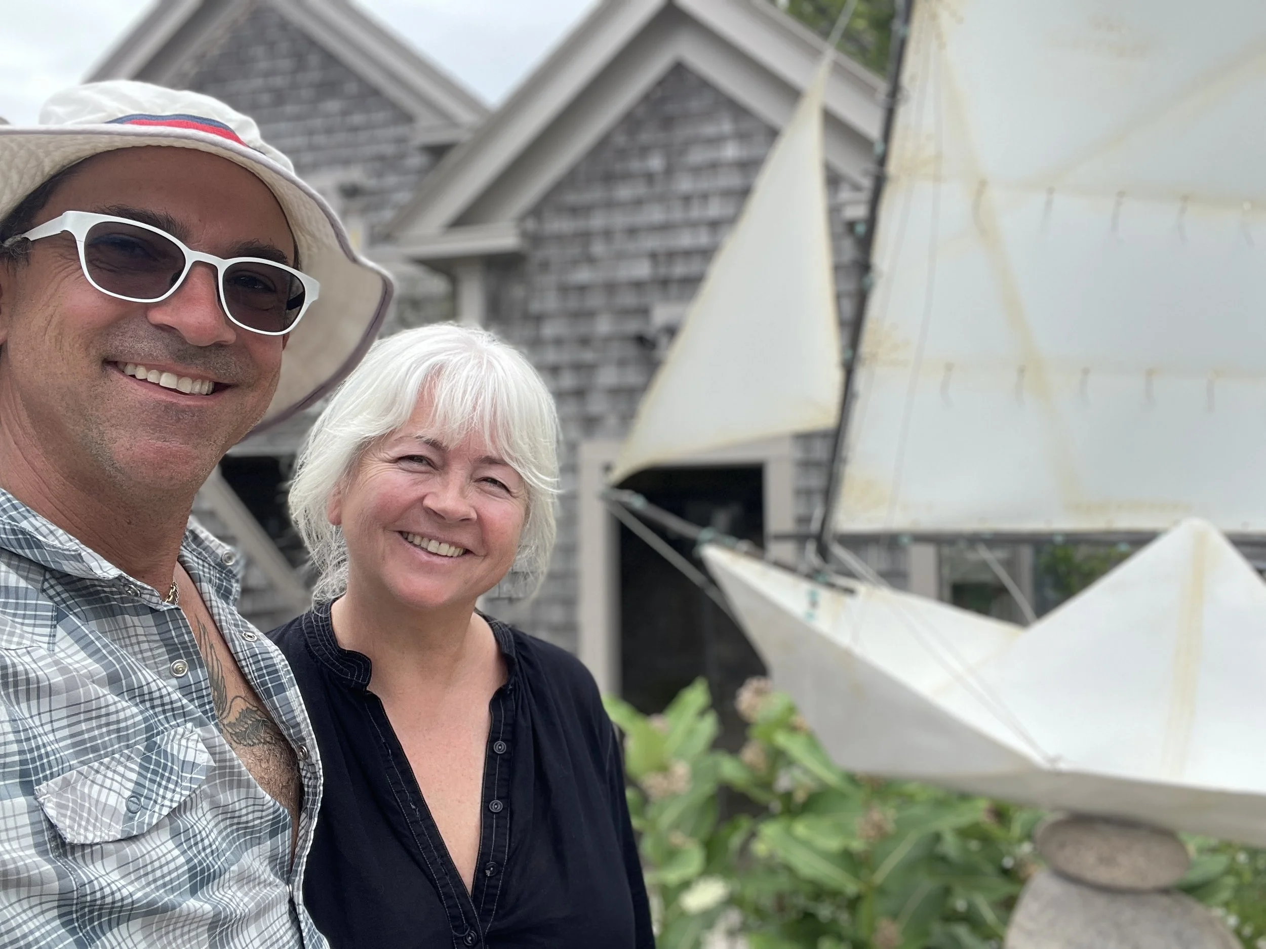Close-up of two smiling women and one smiling man standing outdoors near a white umbrella and a wooden house.
