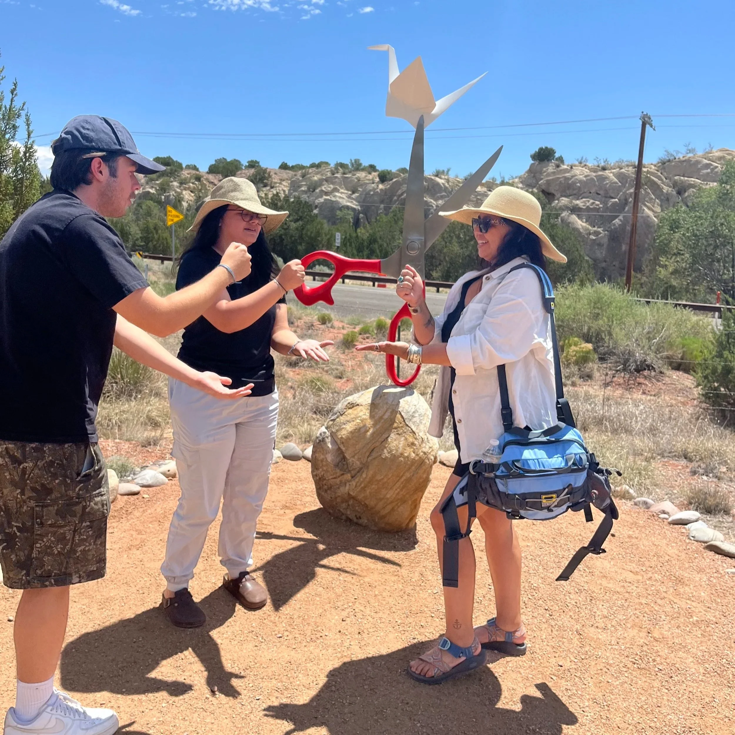 Three women and one man outdoors, holding and comparing a large pair of red scissors, with a sculpture of a bird in the background, on a sunny day with blue skies and rocky hills.