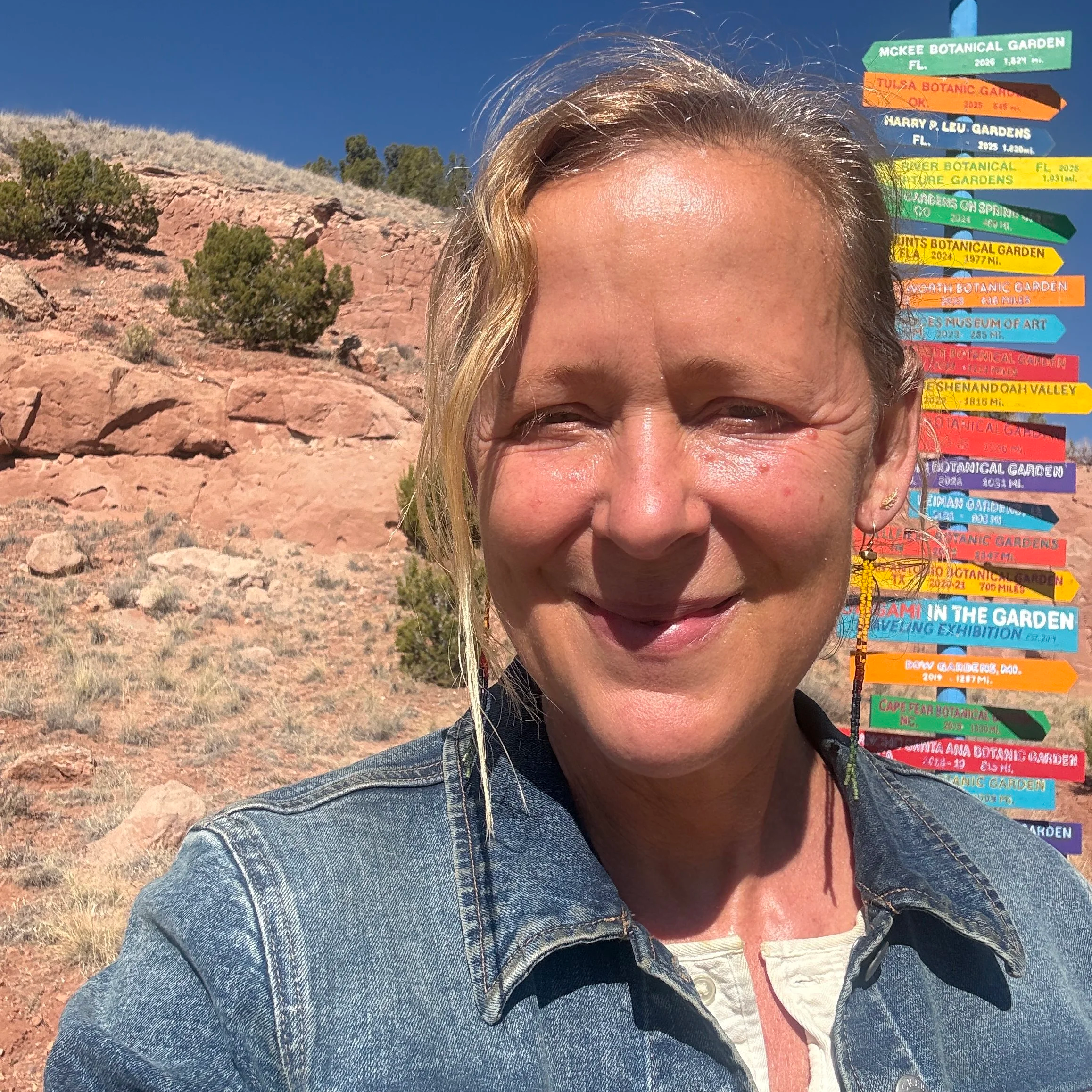 Close-up of a woman smiling outdoors with rocky terrain, green bushes, and a bright blue sky in the background. She is wearing a denim jacket and earrings, and standing next to a colorful directional sign at a botanical garden.