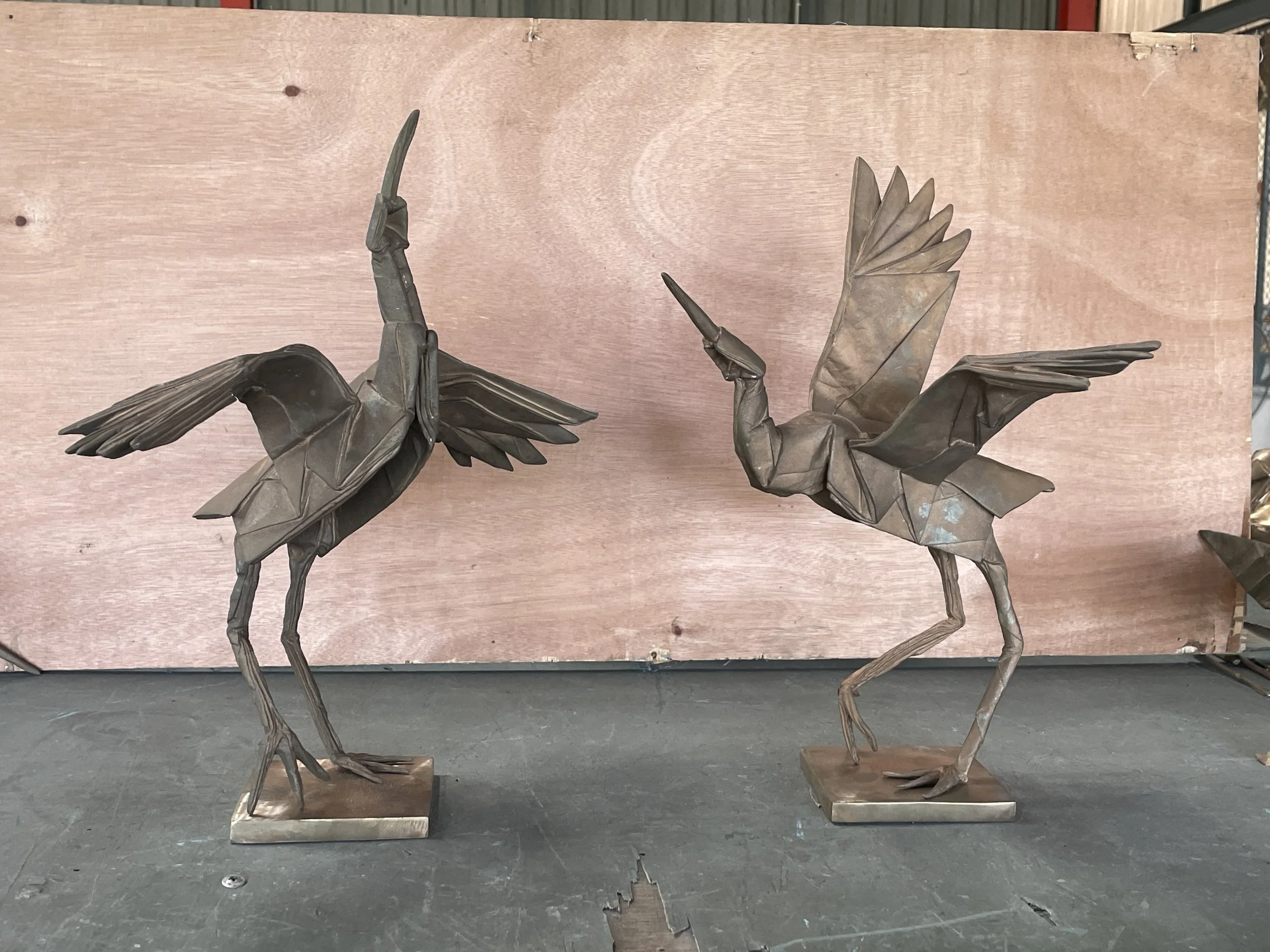 Two metal origami cranes with wings spread, mounted on metal bases, positioned on a black surface against a plain wood-colored background.