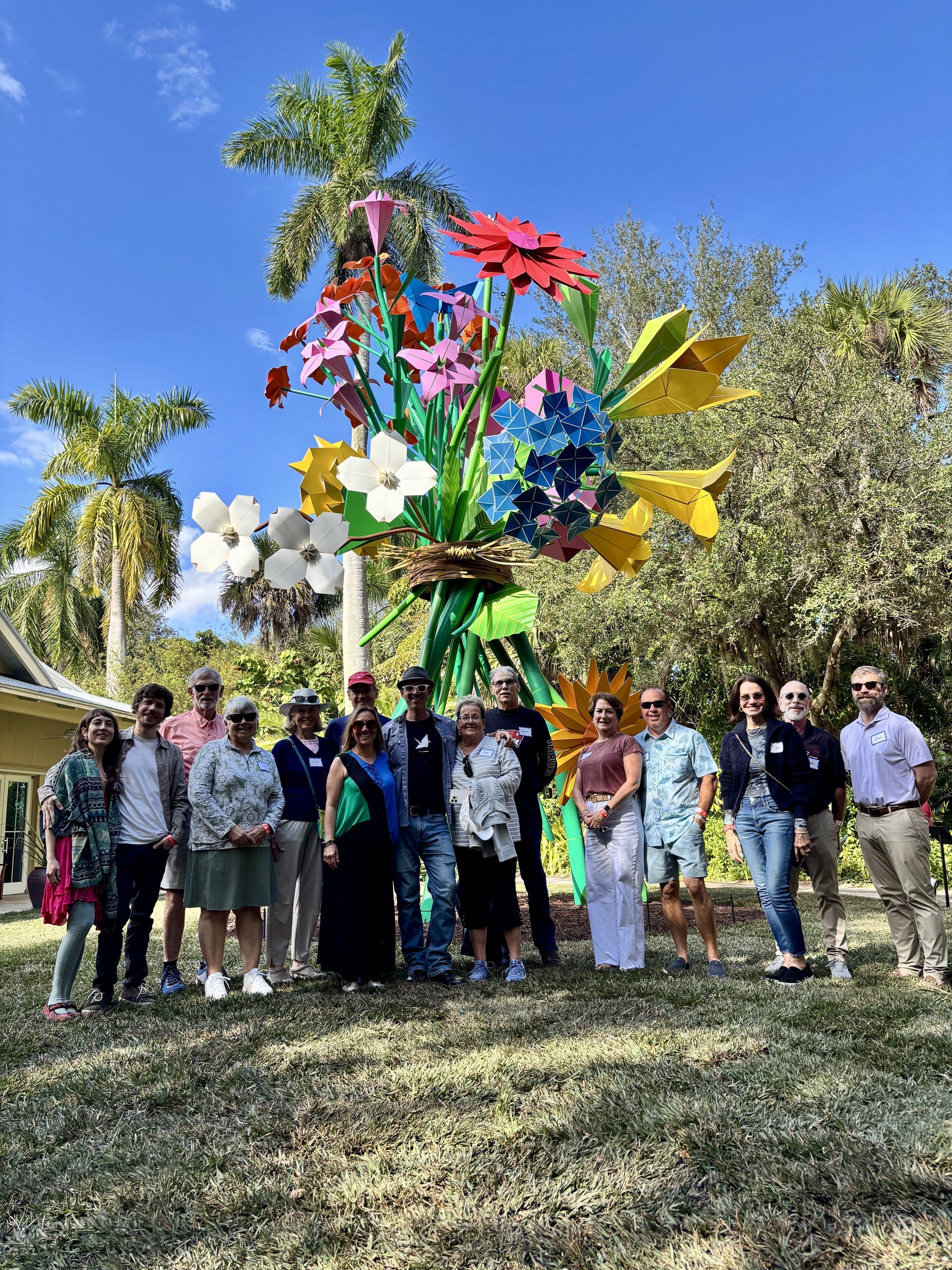 A group of people standing outdoors in front of a colorful, sculpture-like art piece shaped like flowers, with palm trees and a blue sky in the background.