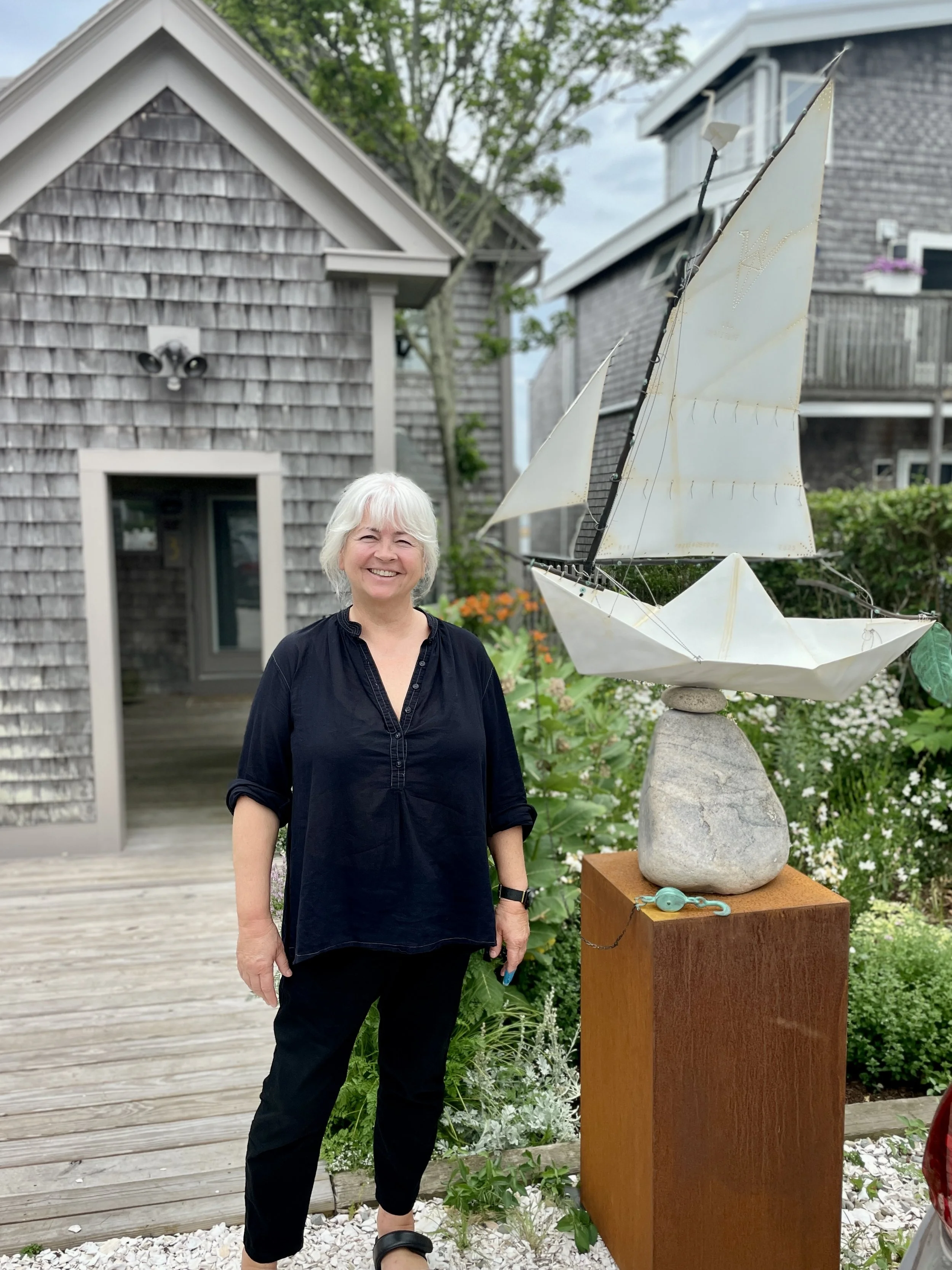A smiling woman with white hair standing outdoors next to a sailboat sculpture made of white material and mounted on a stone base, with residential houses and greenery in the background.