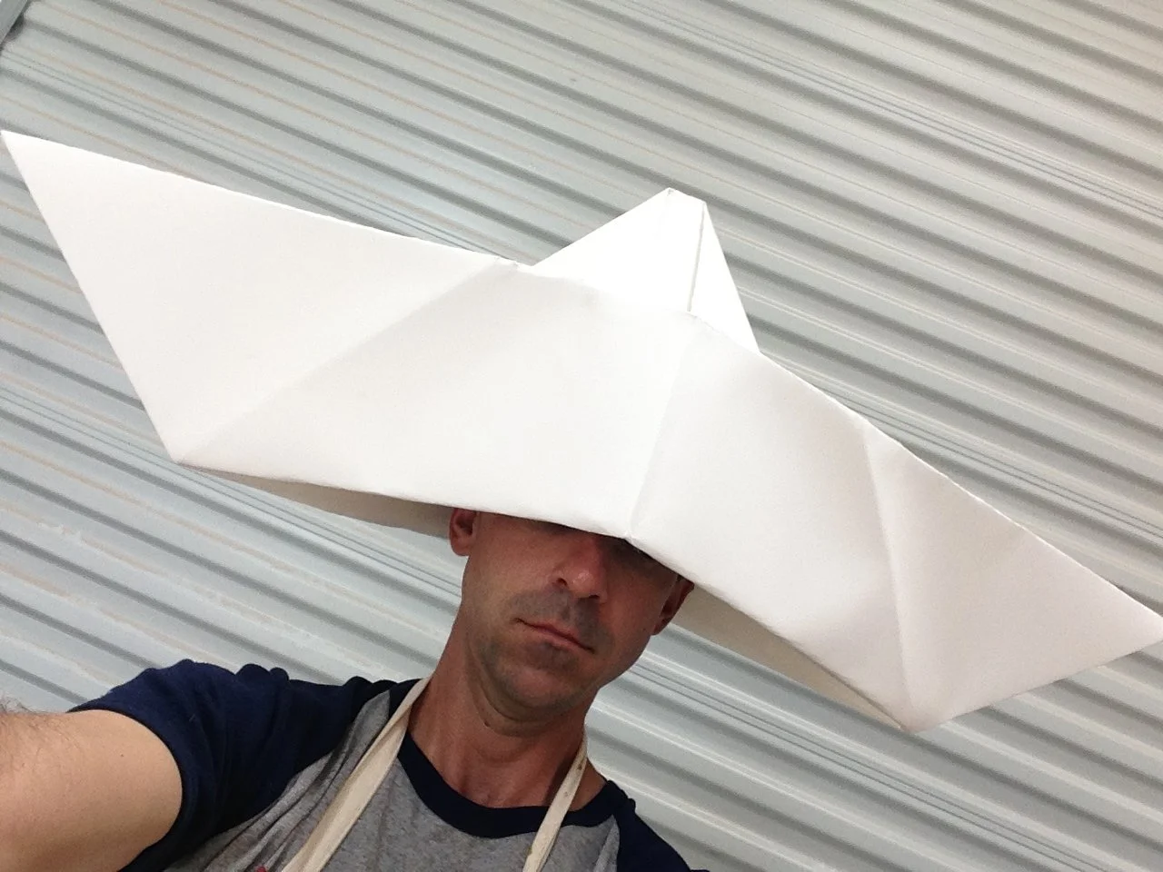 A man wears a large white paper airplane hat covering his head and eyes, standing in front of a metallic roll-up door.