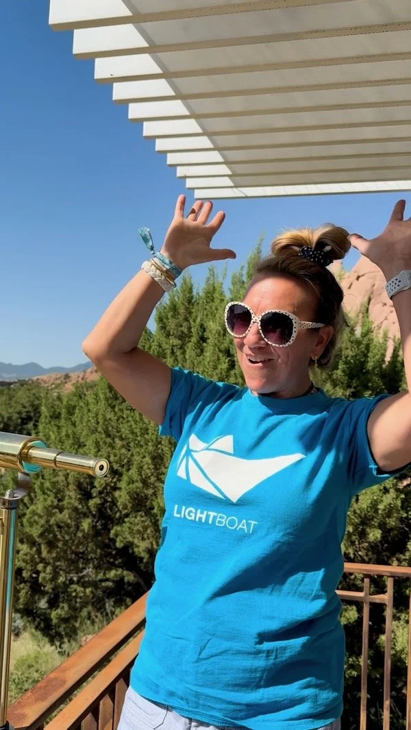 Jennifer Box, wearing a  blue T-shirt featuring an origami boat, stands outdoors on the portico of Box Studio Sculpture Garden with trees and mountains in the background.