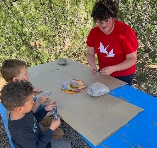 A woman with curly hair wearing a red shirt with sailboats on it is showing rocks to three children sitting at a table outside. The children are holding rocks and looking at the woman, with a table covered in large rocks and other items. The background features trees and greenery.