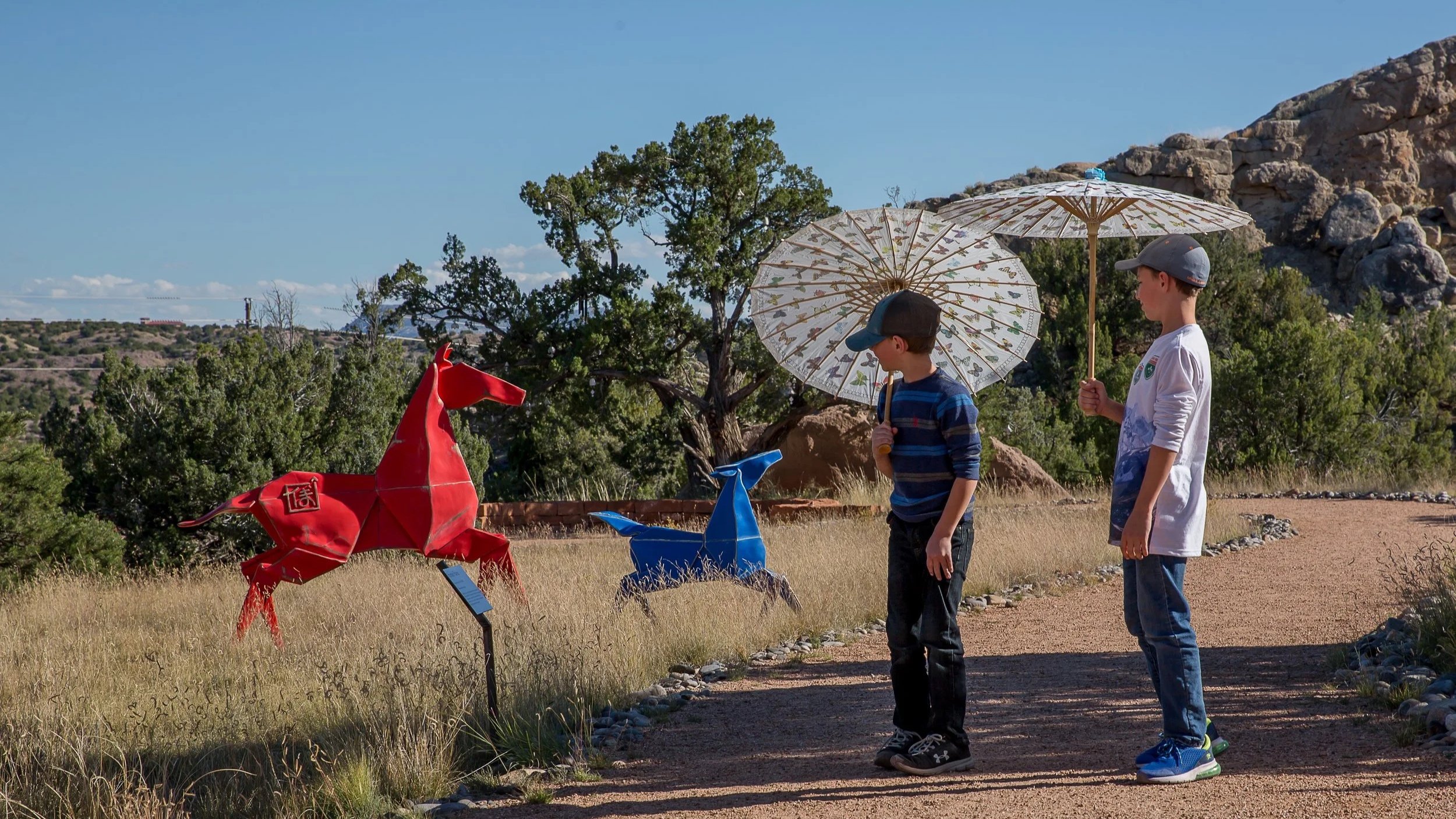 Two boys walk outdoors holding umbrellas, near colorful horse sculptures, in a natural setting with trees and rocky terrain.