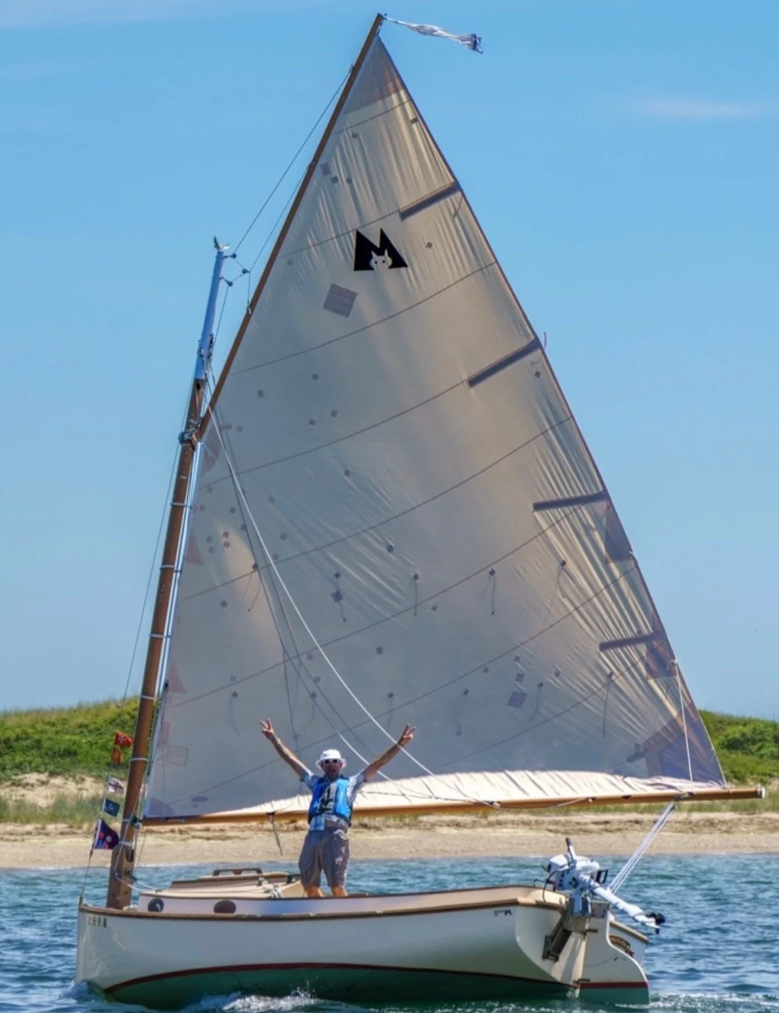 A person stands on a small sailboat with arms raised, on the water near a beach with green hills in the background. The sailboat has a large, shiny sail and a small outboard motor.