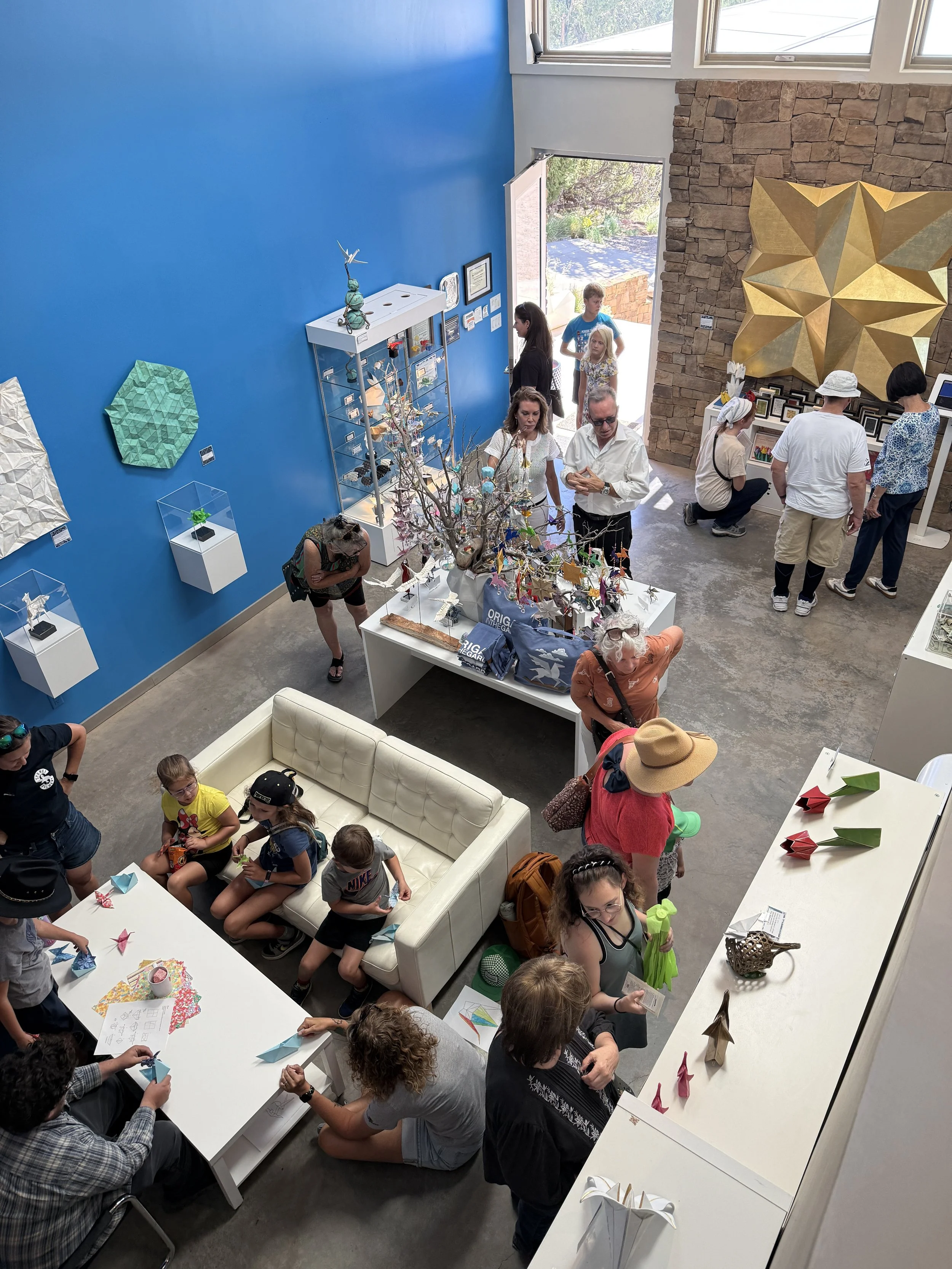 Visitors gather in the gift shop and gallery at Box Studio Sculpture Garden in Cerrillos, New Mexico, featuring the Inside Out exhibit of paper origami models and cast metal unfolded origami on blue walls.