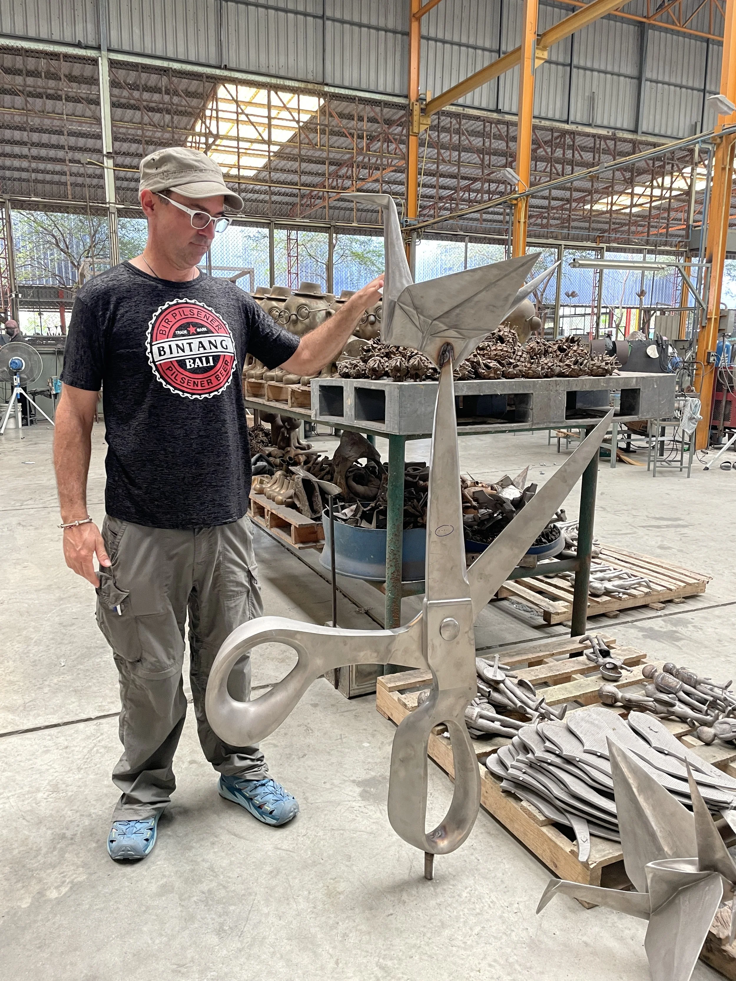 A man wearing glasses, a cap, a black T-shirt, and gray pants is inspecting a large metallic sculpture of a snake in a workshop. The workshop contains various metal parts and sculptures on tables and pallets, with an industrial setting including a high ceiling, metal beams, and large windows.