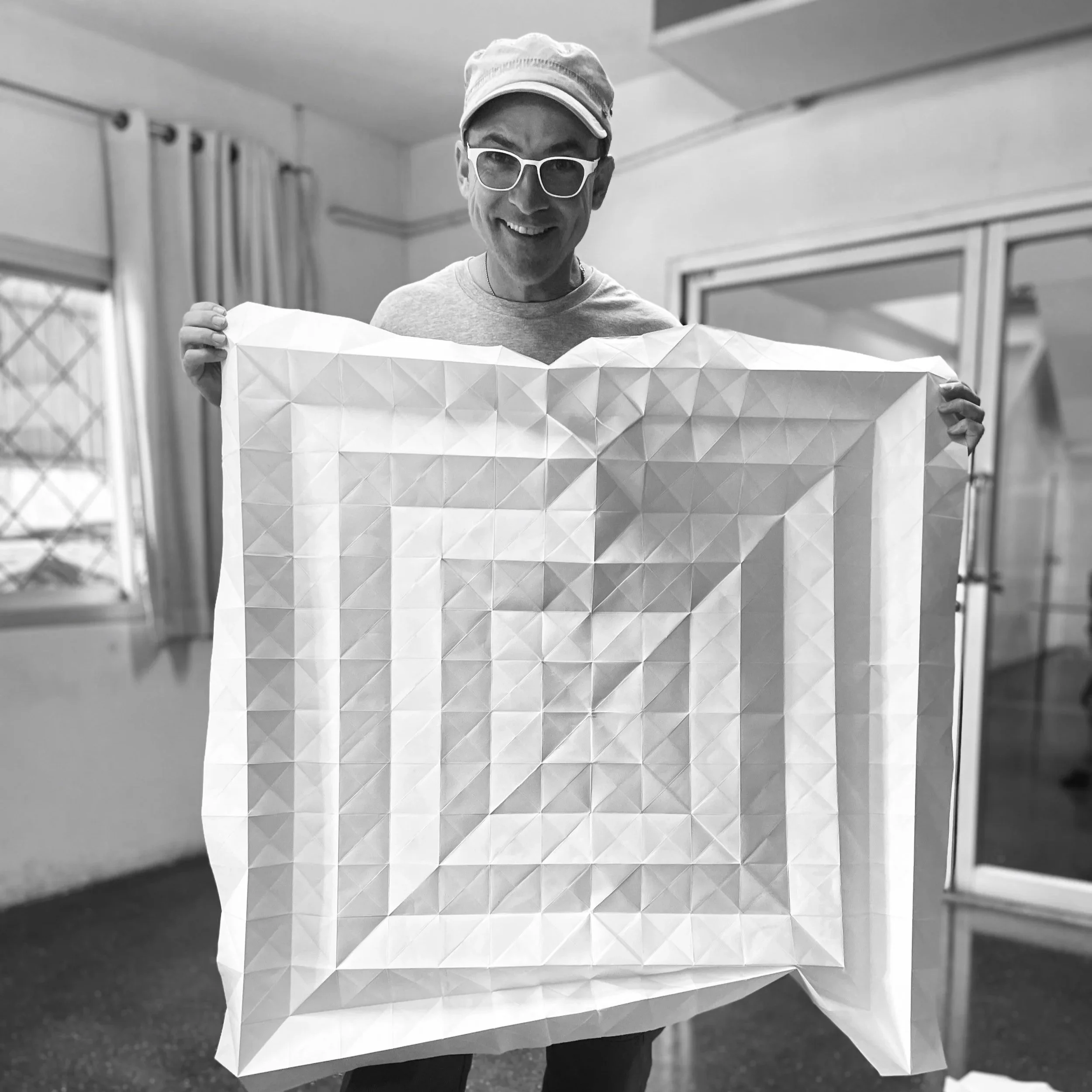 A man wearing glasses and a hat smiles while holding a geometric patterned sheet. The background shows a room with a window and curtains.
