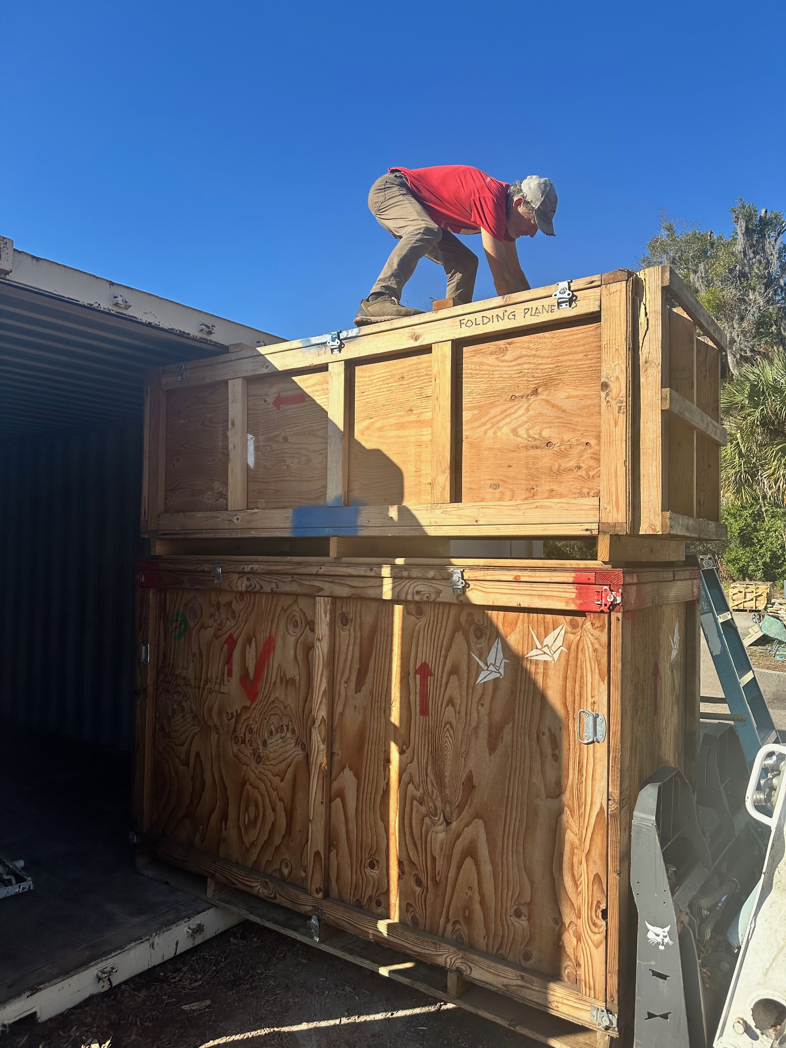 A man in a red shirt, beige pants, and a gray hat is working on the top of a wooden structure labeled "Folding Plane." The structure is partially assembled and is attached to a vehicle, with some metal hinges visible. The background shows trees and a clear blue sky.