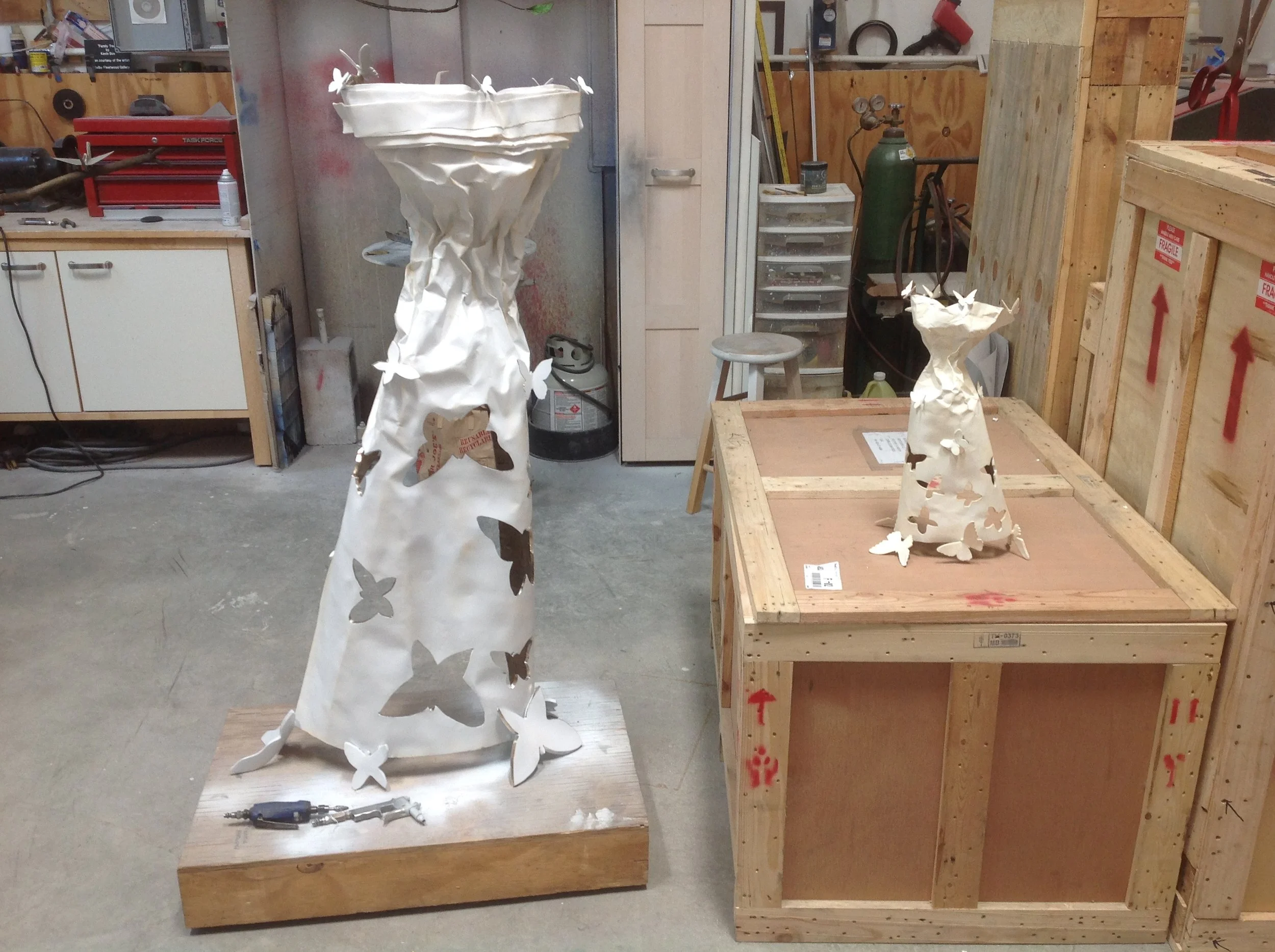 Two decorative trees made of white material with butterfly cutouts, placed on wooden bases in a workshop.