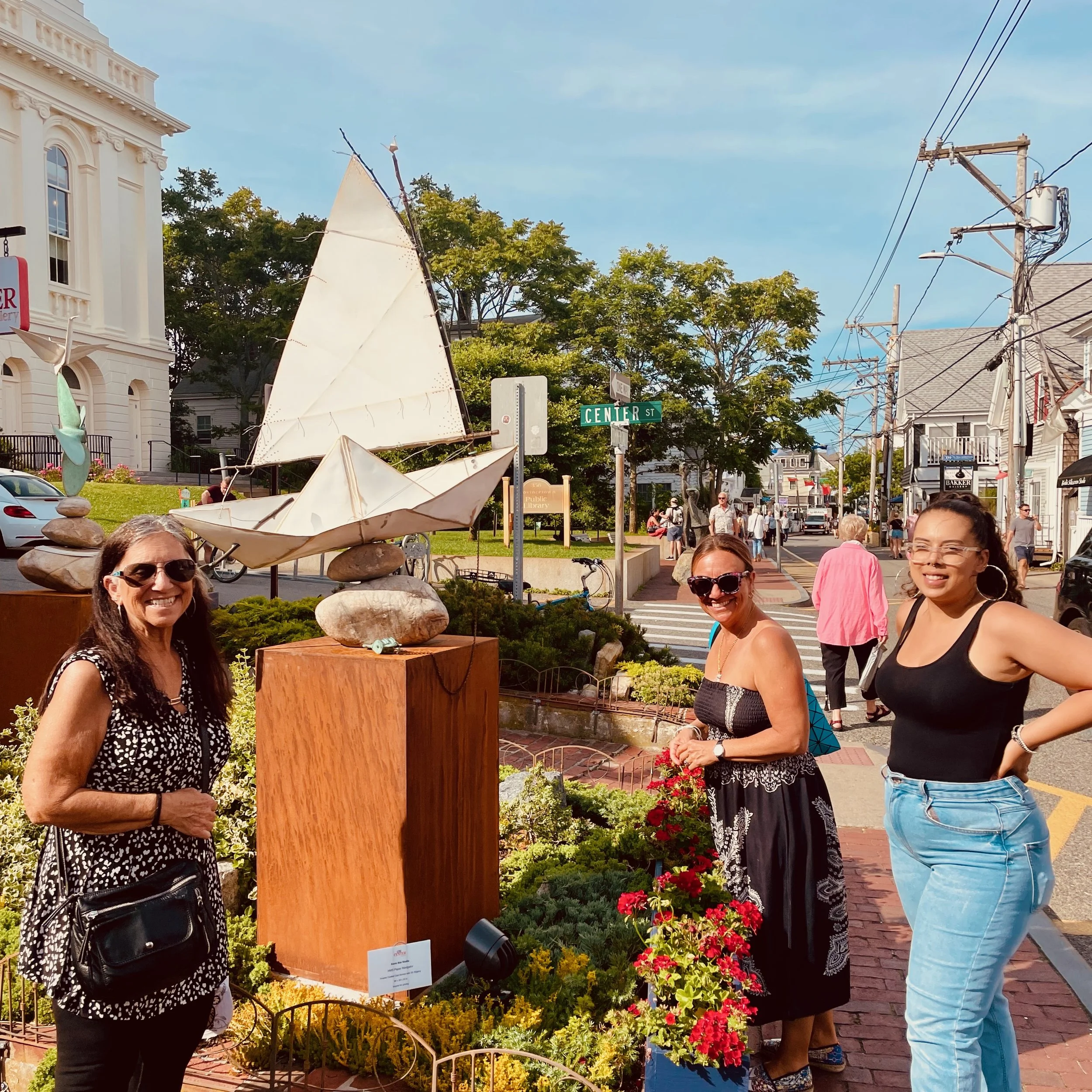 A street scene on Center Street with a group of women smiling, a white building on the left, a small sailboat sculpture in the foreground, and power lines overhead. There are many people walking and shopping on the sidewalk.