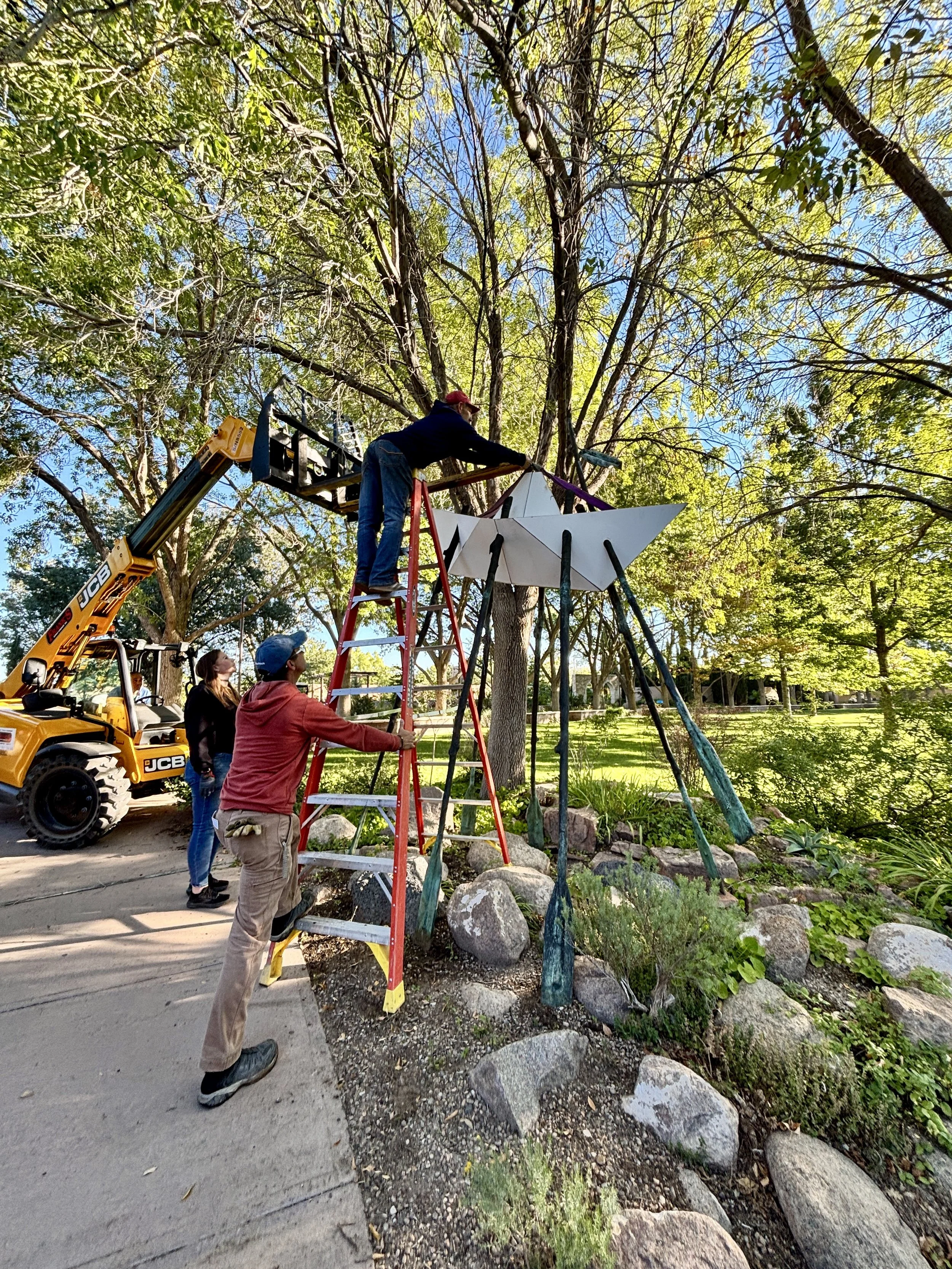 Garden teams install a tall sculpture of a white metal paper boat on oars using heavy machinery at the ABQ BioPark Botanic Garden