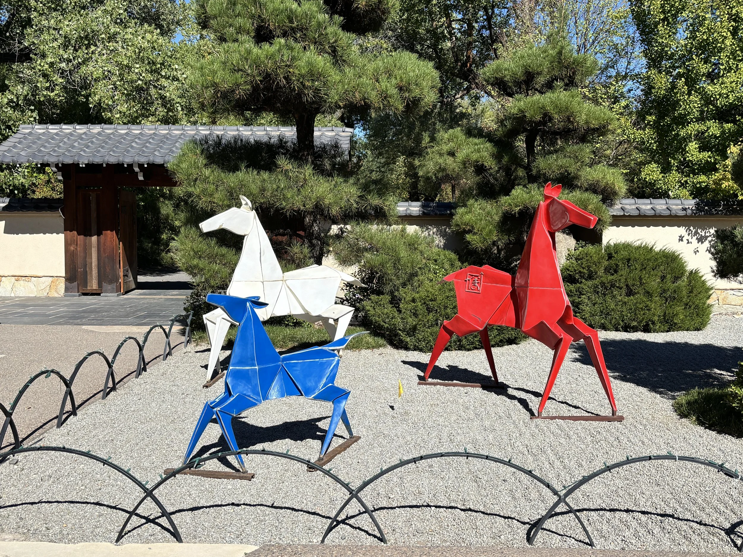 Three metal origami ponies in red, white and blue stand in the raked rock bed of the Japanese Garden at ABQ BioPark Botanic Garden.