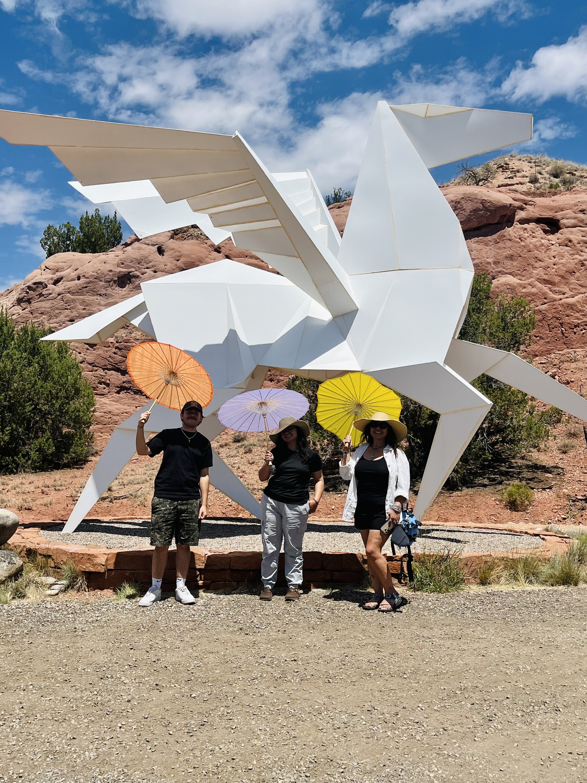 Three people standing in front of a large white geometric sculpture resembling a dragon, each holding colorful parasols, in a desert landscape with red rocks and a blue sky with clouds.