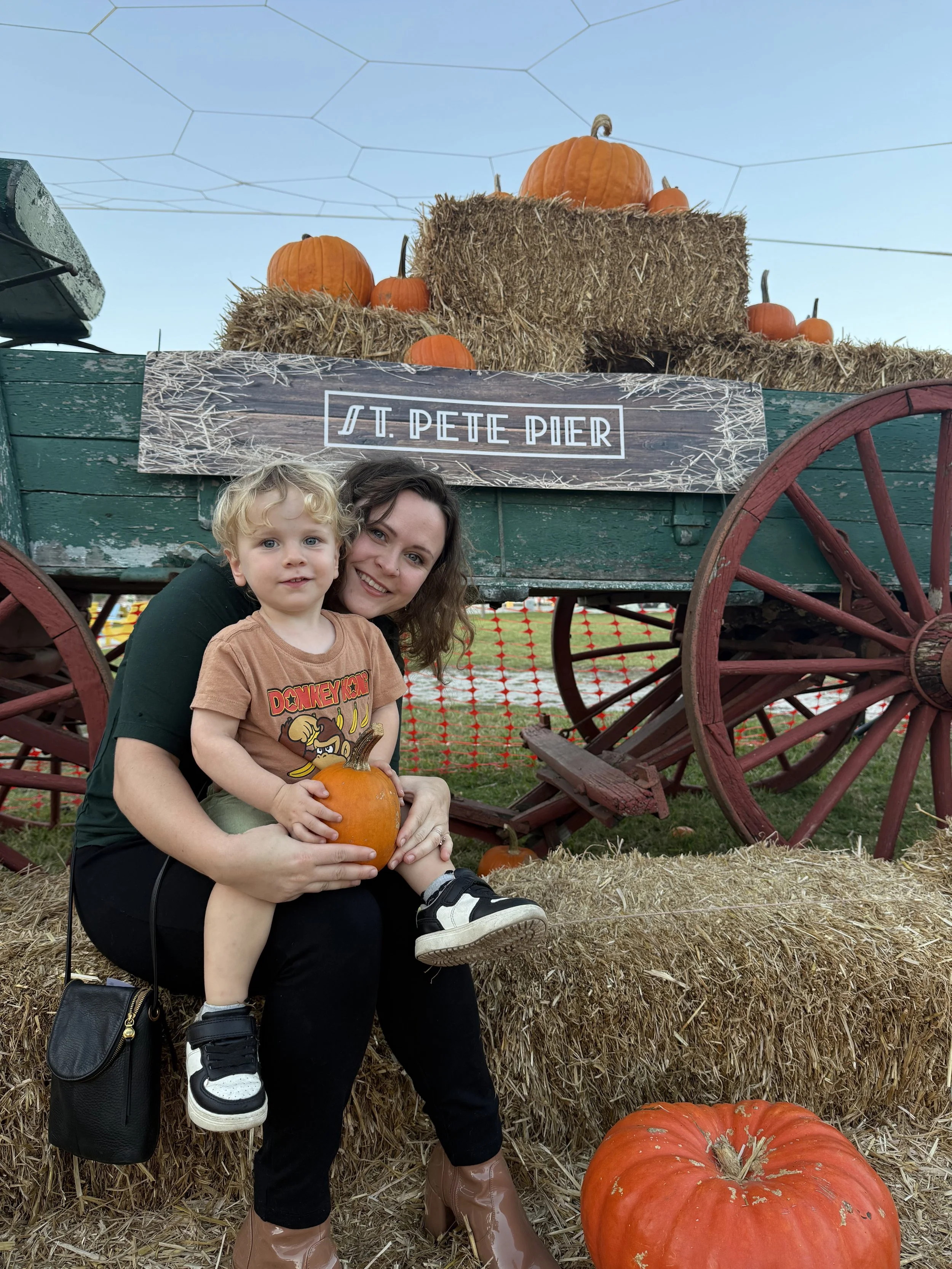 A woman and a young boy sitting on hay bales at a pumpkin patch, with a decorated wagon in the background with pumpkins and hay, and a sign that reads 'St. Pete Pier'. Photographer videographer St. Petersburg Florida St Pete Saint Petersburg