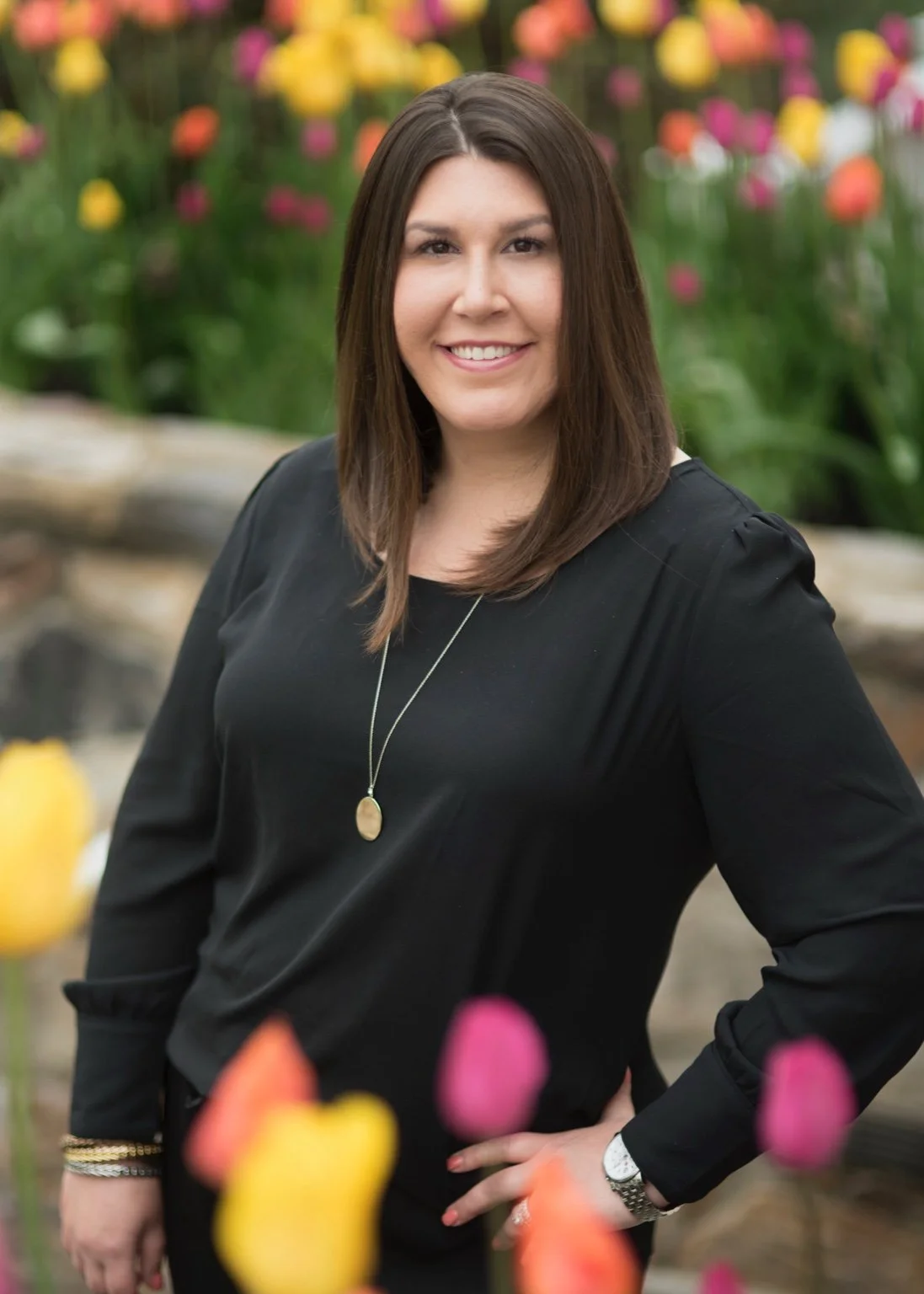 A woman with brown hair smiling outdoors in front of colorful tulips, wearing a black dress and a long pendant necklace.