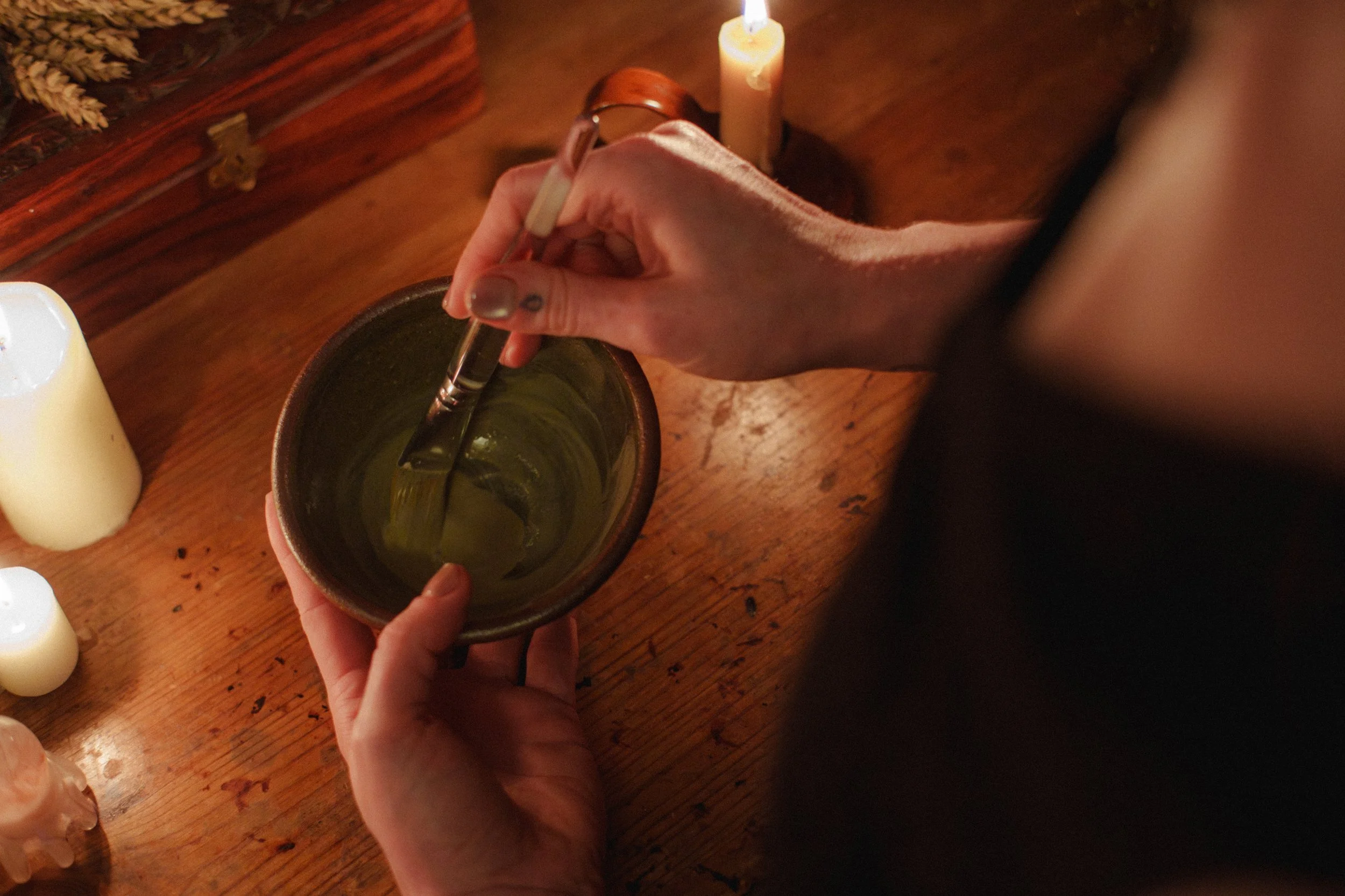 A person holding a small ceramic bowl filled with a face mask, with a candle and some objects on a wooden table in the background