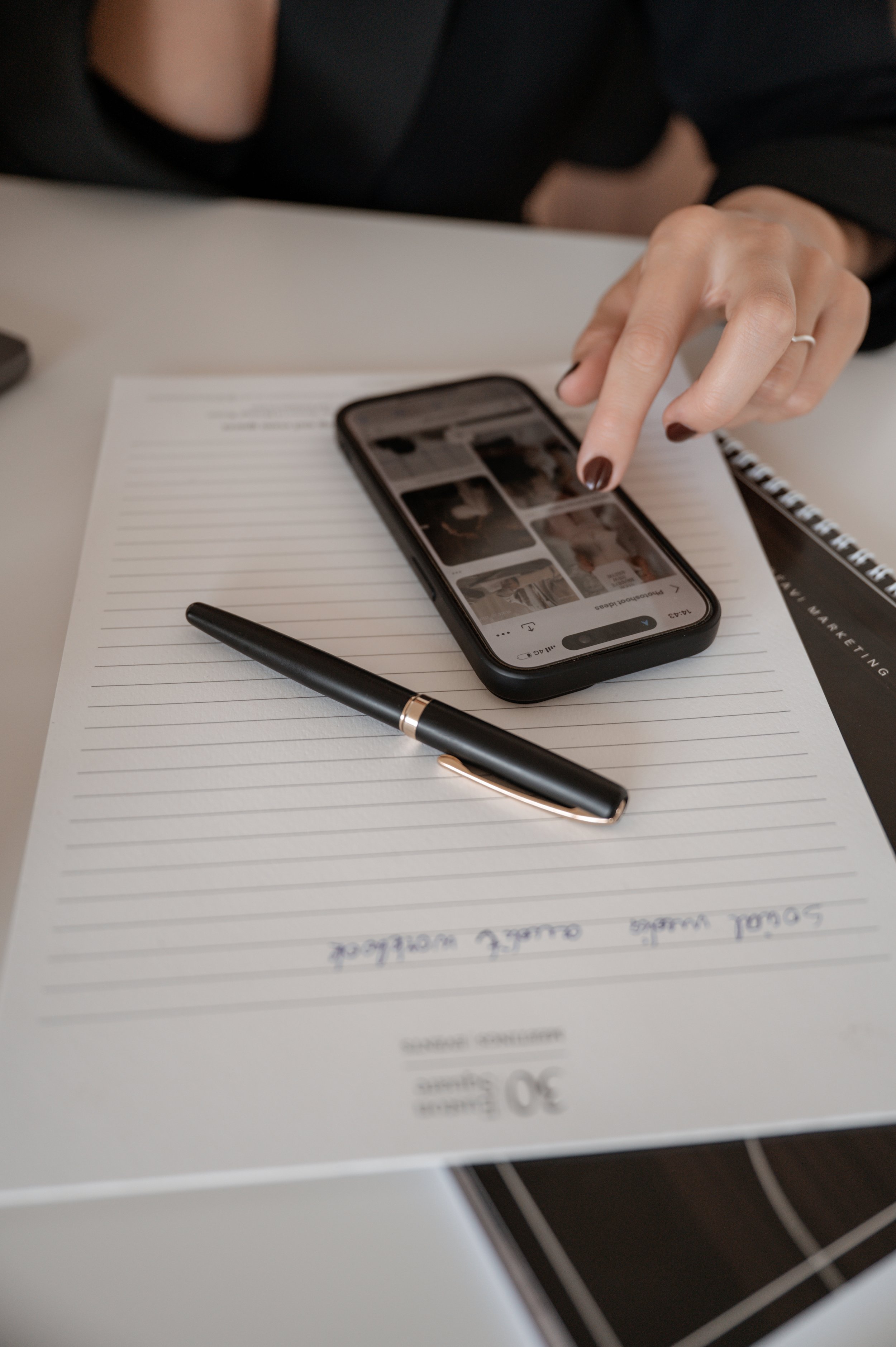 Close-up of a person's hand with dark nail polish pointing at a phone displaying a photo gallery, on top of a white lined notepad with handwritten notes, a black pen, and a spiral-bound book, on a desk.