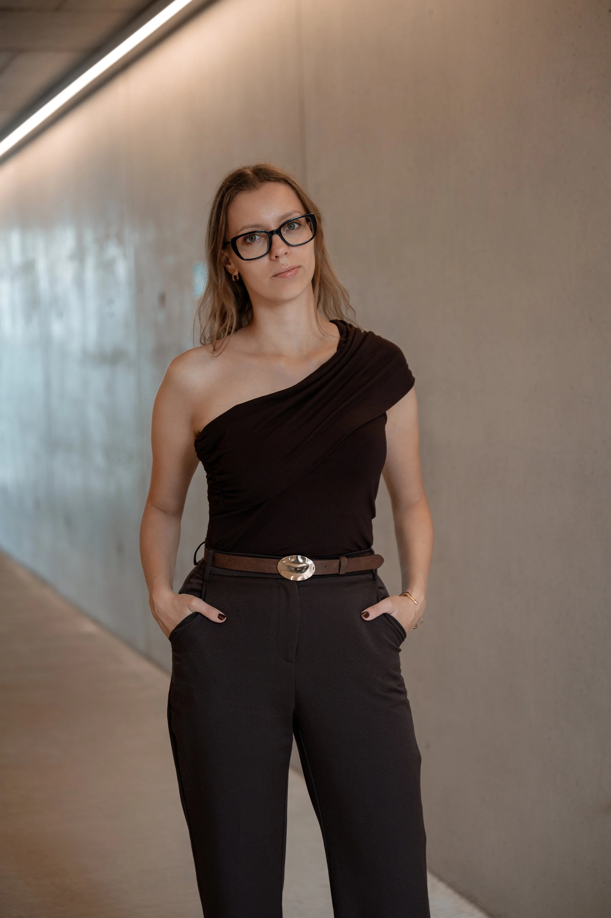 Woman with red hair and glasses standing in a hallway with her hands in her pockets, wearing a black one-shoulder top and black pants.