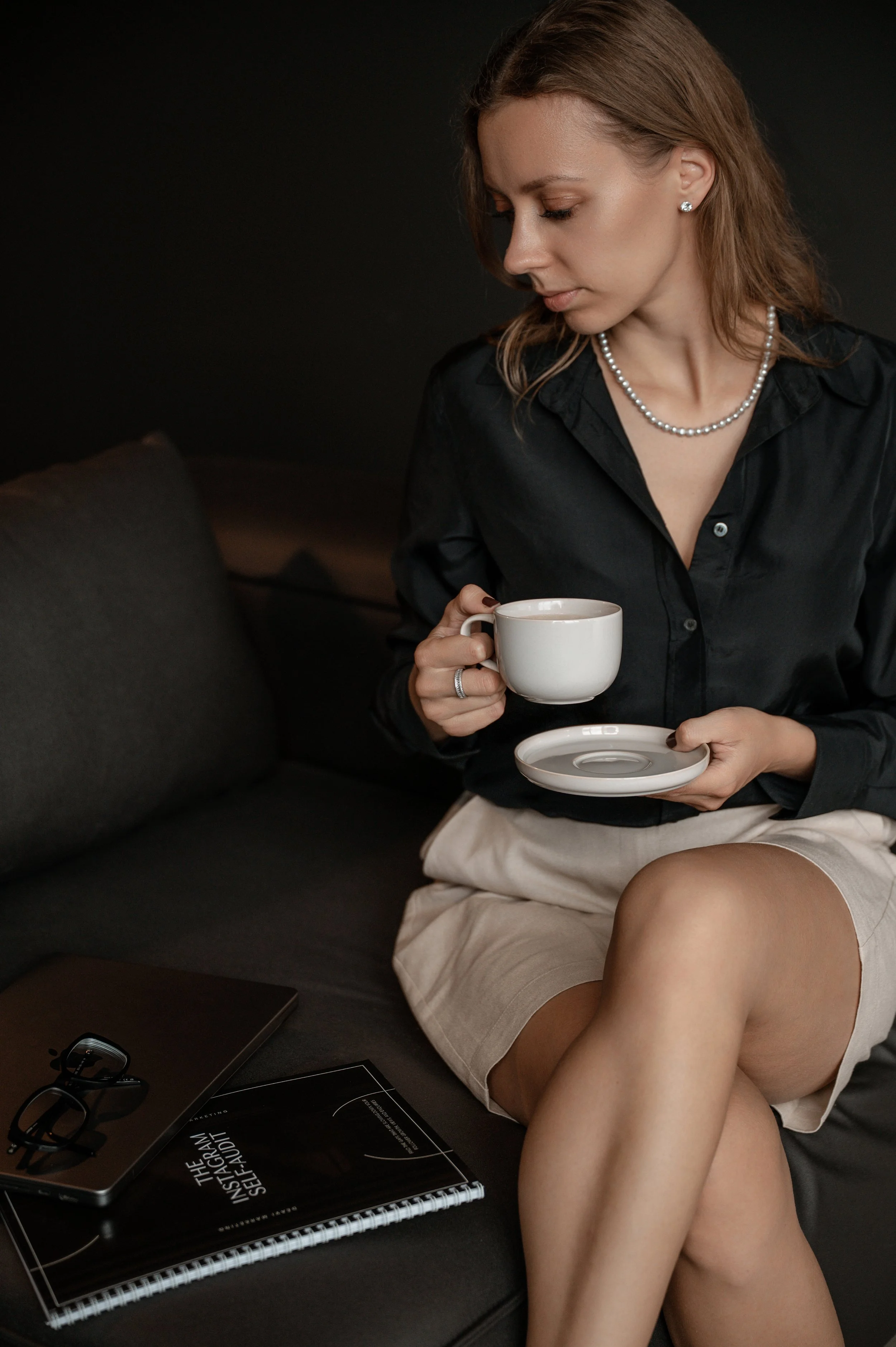 A woman with brown hair, wearing a black shirt, pearl necklace and earrings, is sitting on a couch holding a white coffee cup with saucer. She is looking down at the cup. There are sunglasses, a black electronic device, a magazine titled 'The Algorithm', and a spiral notebook on a black table beside her.