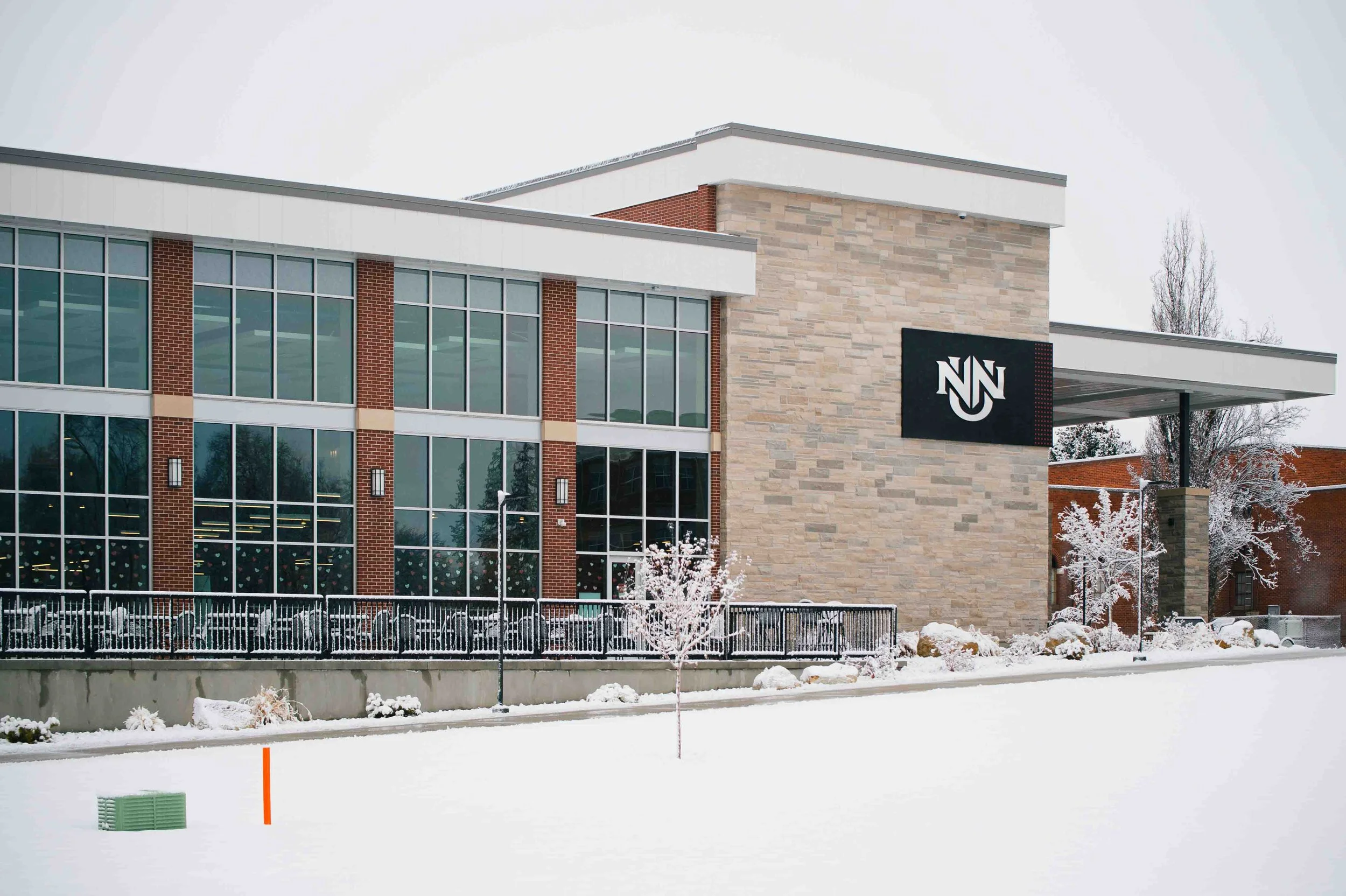 Modern building with large glass windows, brick and stone exterior, black signage with white letters, surrounded by snow and leafless trees in winter.