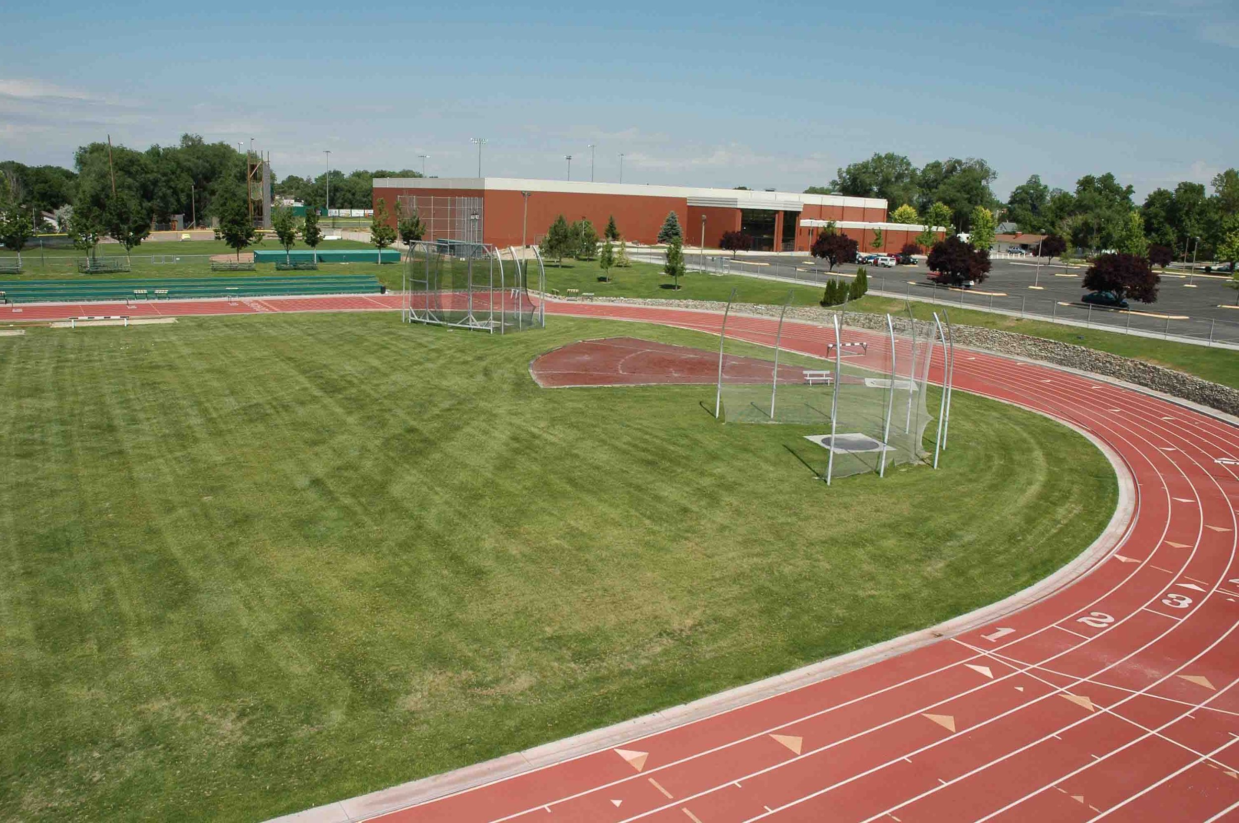 An outdoor sports complex with a running track surrounding a grassy field, with poles and nets for discus and shot put, and a brick building in the background. There are parking lots and trees around the area.