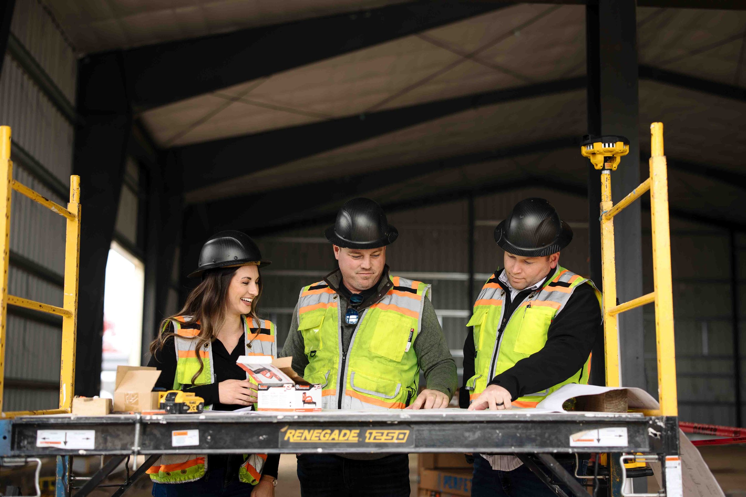 Three construction workers, two men and one woman wearing safety vests and helmets, standing at a work table inside a warehouse, reviewing documents and a toolbox.