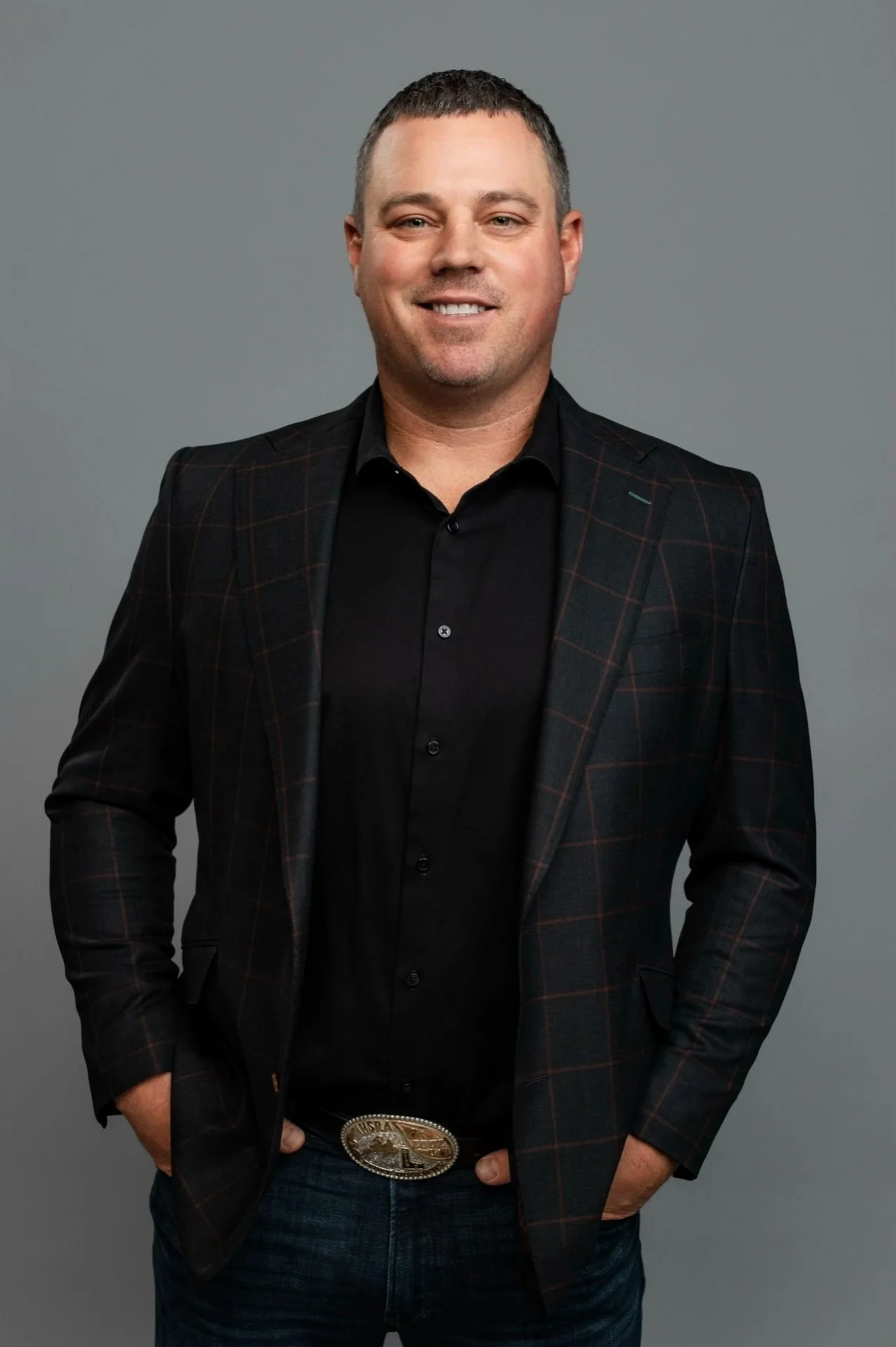A man in a black shirt and plaid blazer with his hands in his pockets, standing against a gray background.