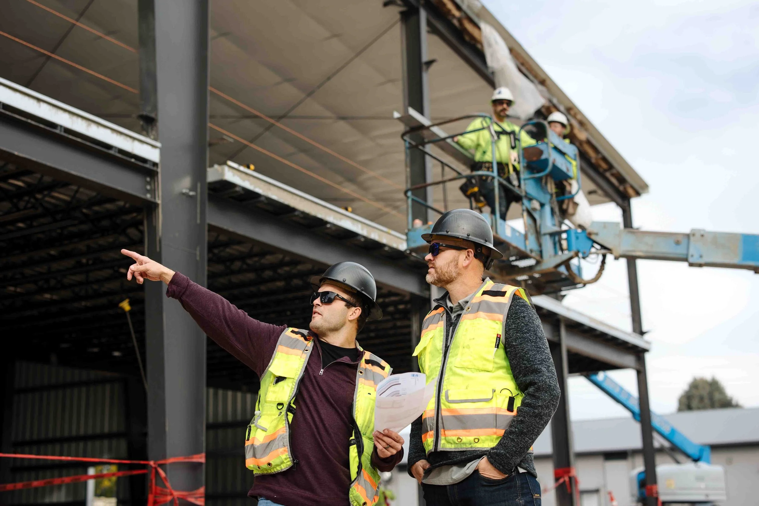 Two construction workers wearing hard hats and safety vests discussing at a construction site, with additional workers on a lift in the background and a building frame under construction.