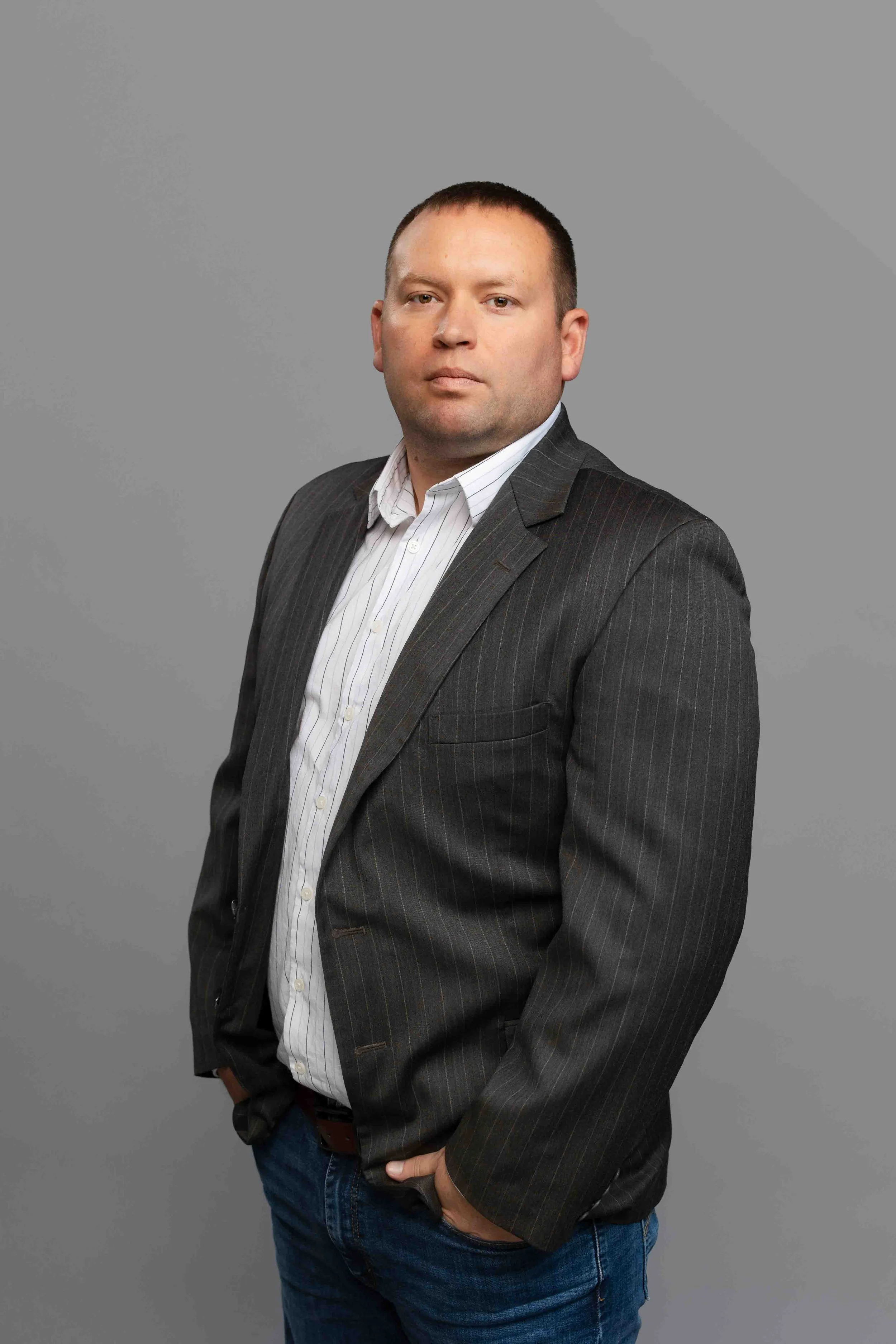 Man in a dark pinstripe blazer and white shirt posing against a plain gray background.