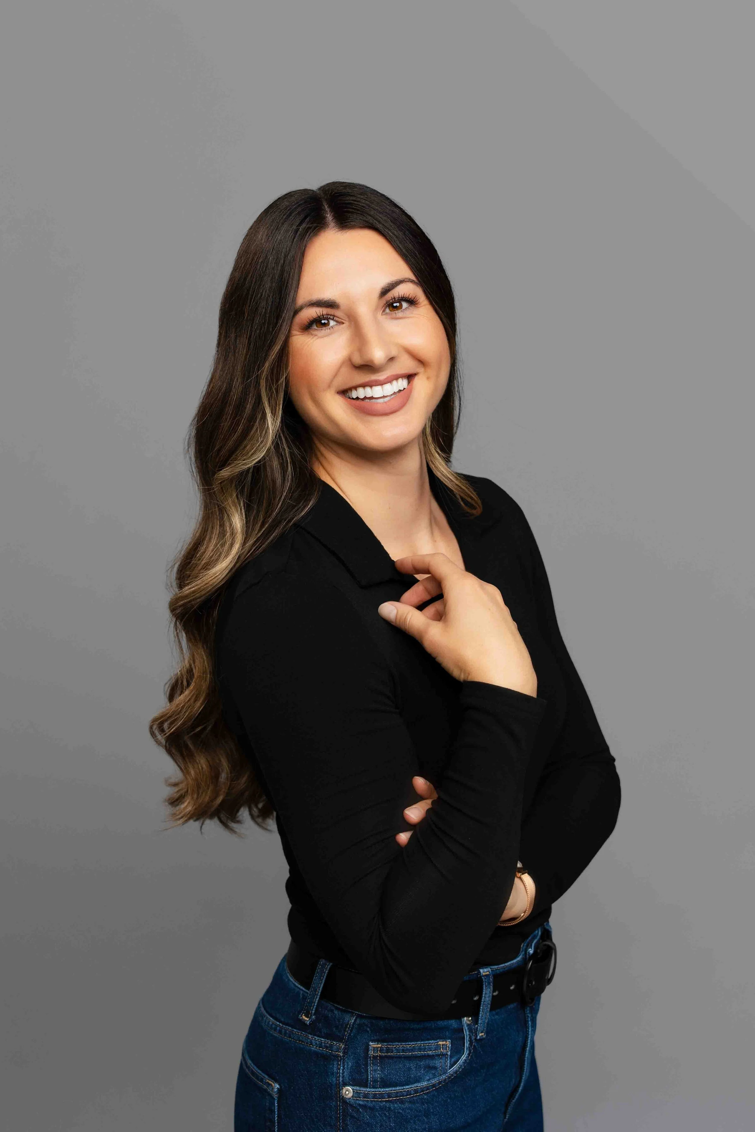 A woman with long wavy brown hair smiling, wearing a black long-sleeve top and blue jeans, standing against a gray background.