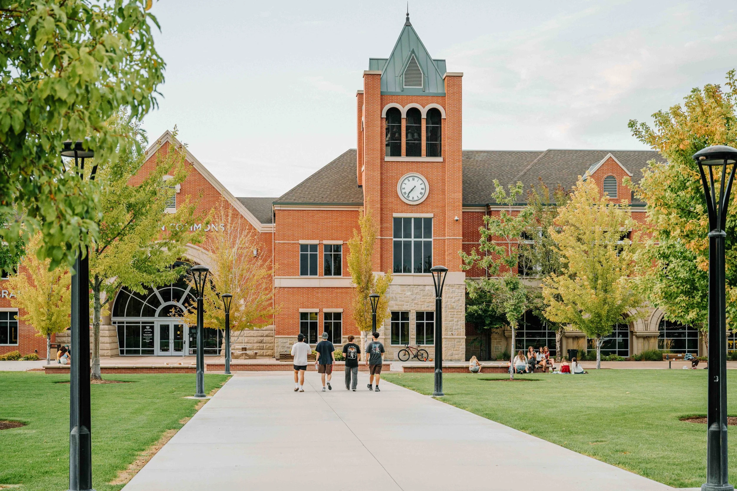 A university campus with a brick building and a clock tower, surrounded by trees, lamp posts, and students walking and sitting on benches.