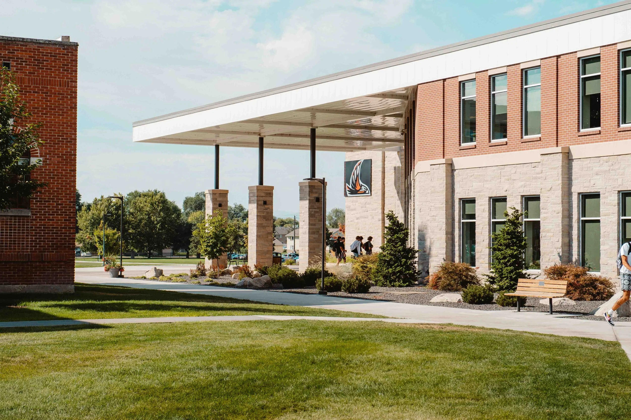 Exterior view of a modern school building with brick and stone facade, large windows, and an overhanging roof supported by columns, with students walking nearby and children in the background.