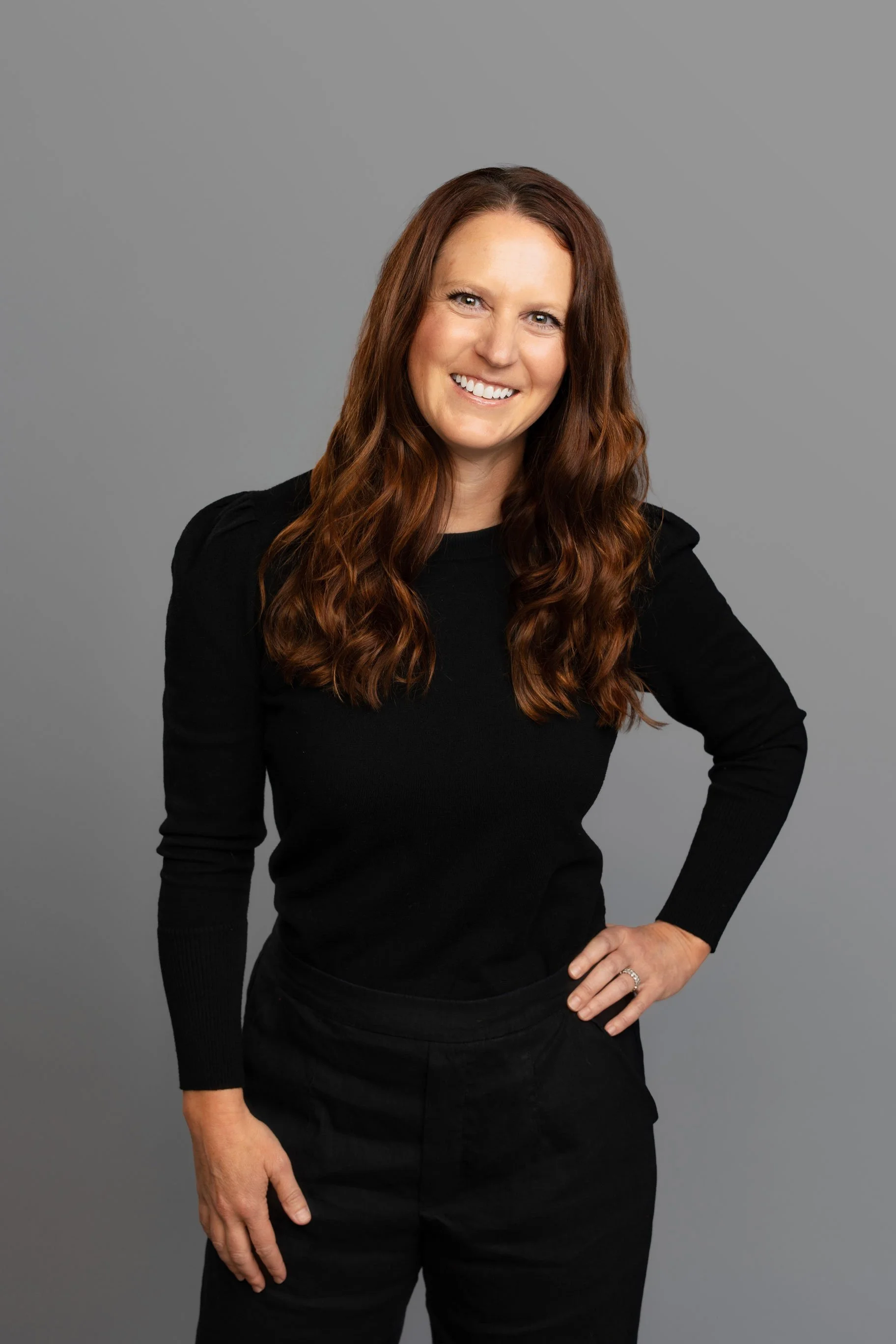 A woman with long wavy red hair smiling and wearing a black long sleeve top and black pants, standing against a plain grey background.