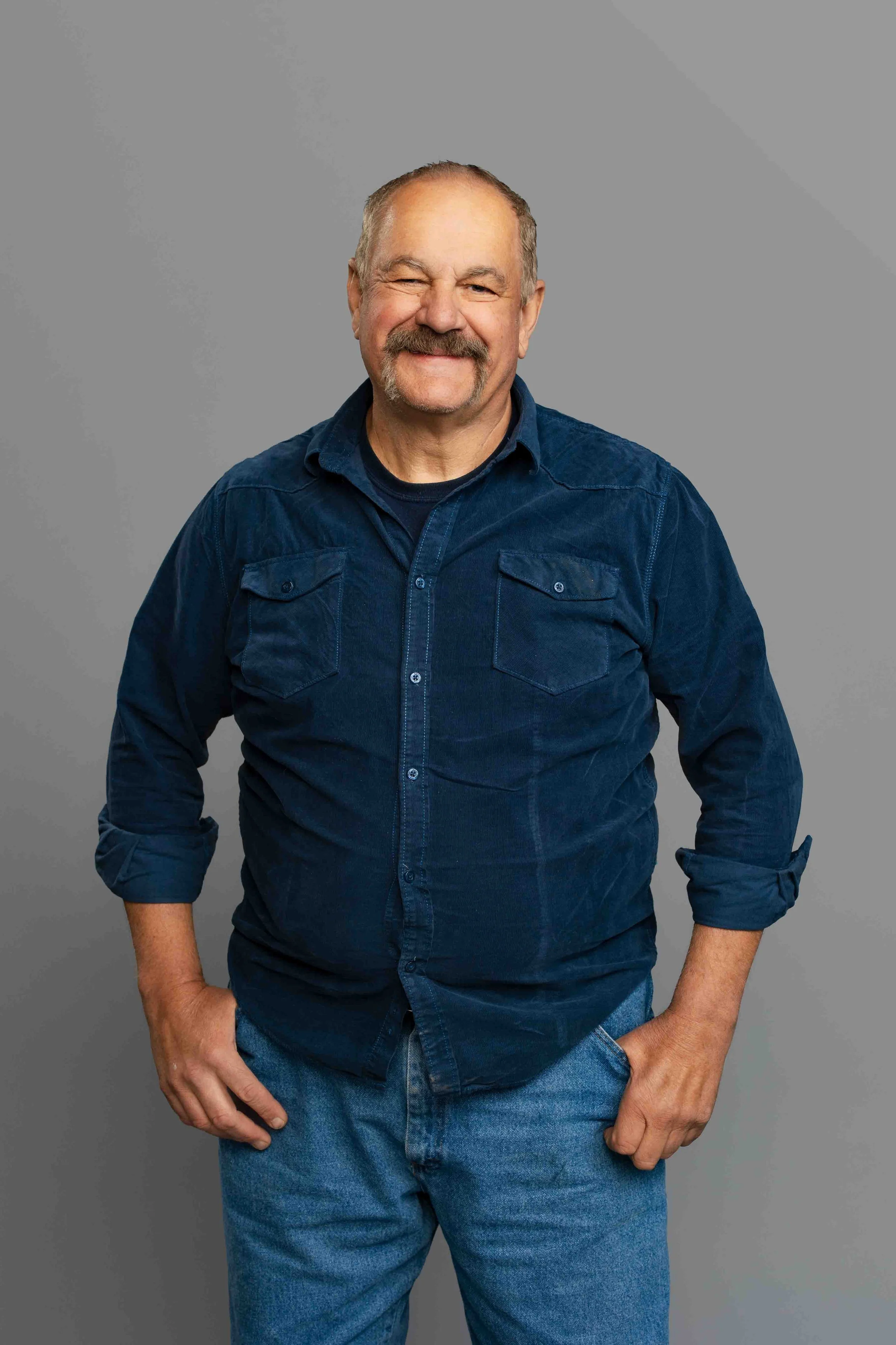 A smiling middle-aged man with a mustache and beard, wearing a blue button-up shirt and jeans, standing against a plain gray background.