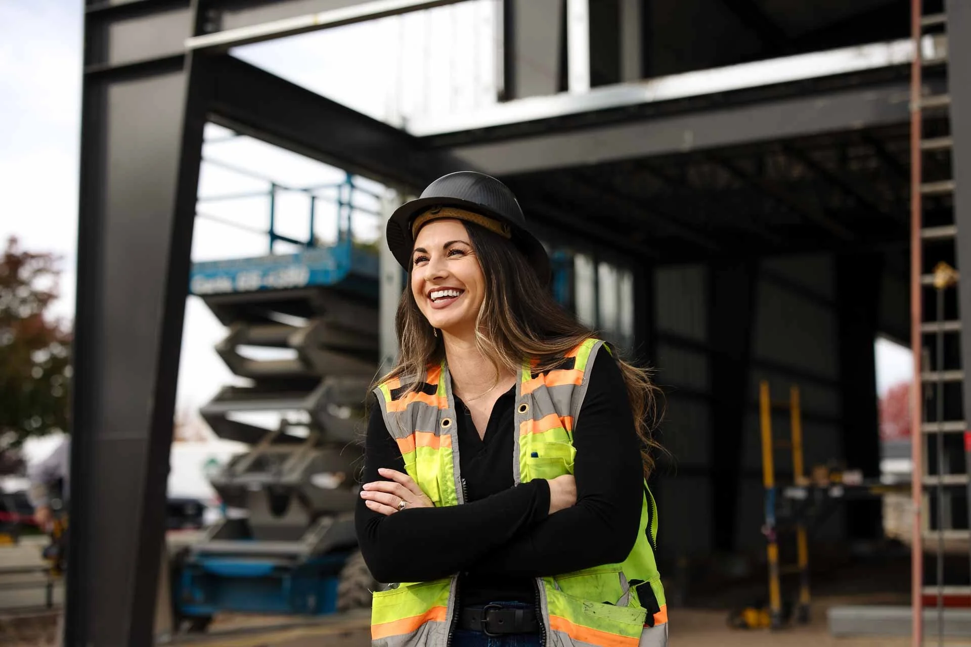 A smiling woman with long brown hair wearing a yellow safety vest and a black hard hat, standing on a construction site with building materials and scaffolding in the background.