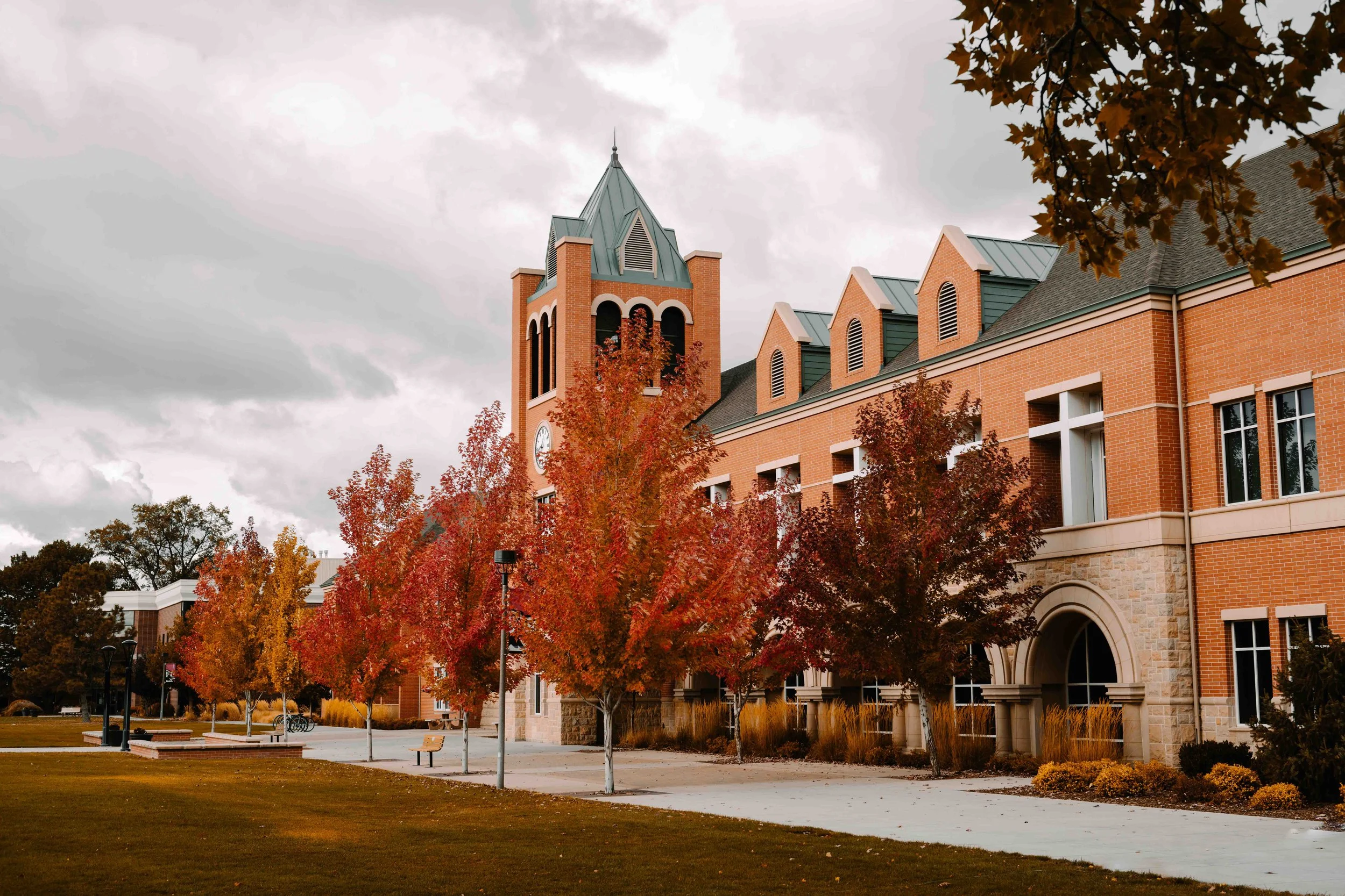 A brick building with a clock tower and green roofs, surrounded by trees with red and orange autumn leaves, on a cloudy day.