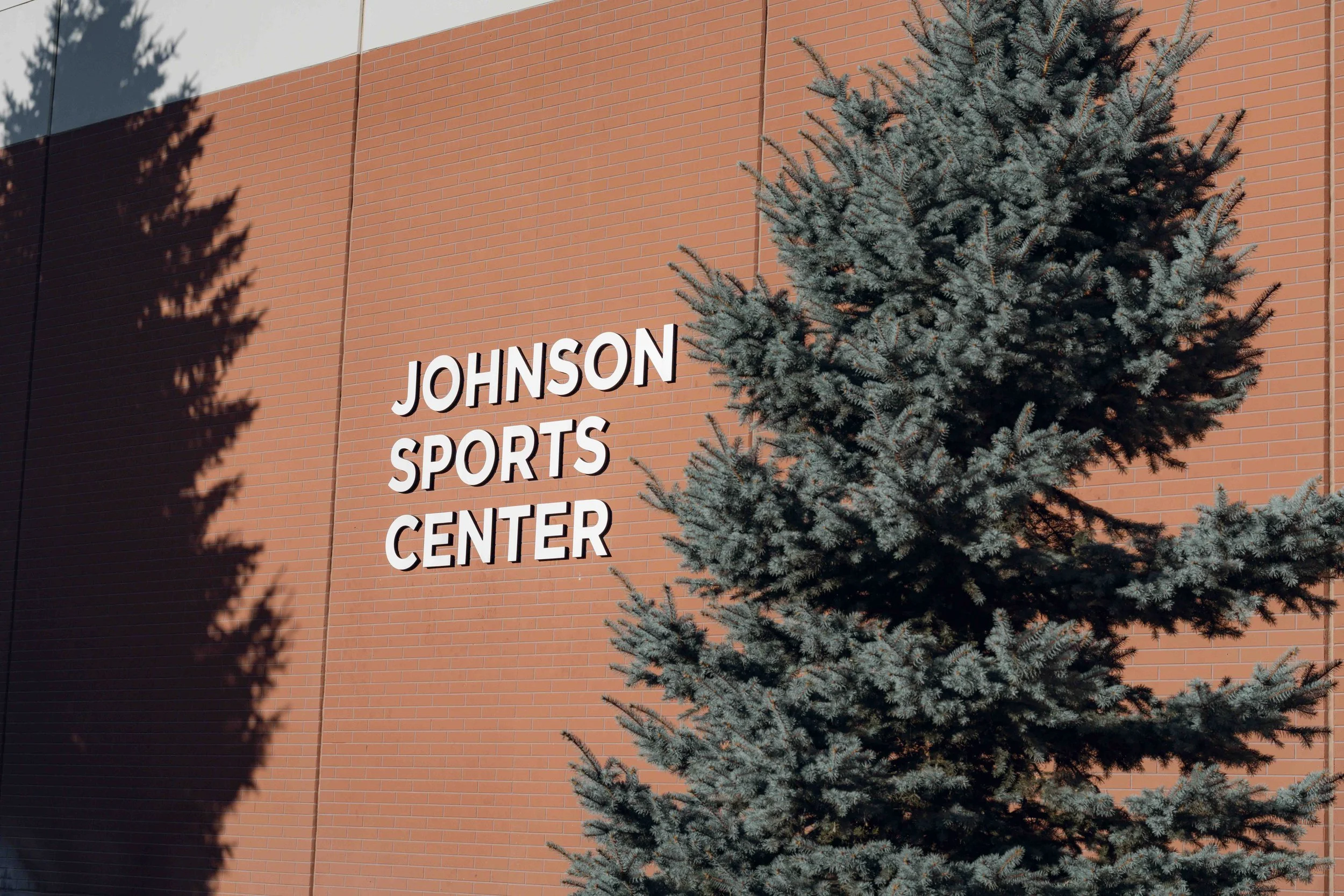 The exterior of the Johnson Sports Center building with a large evergreen tree in the foreground.