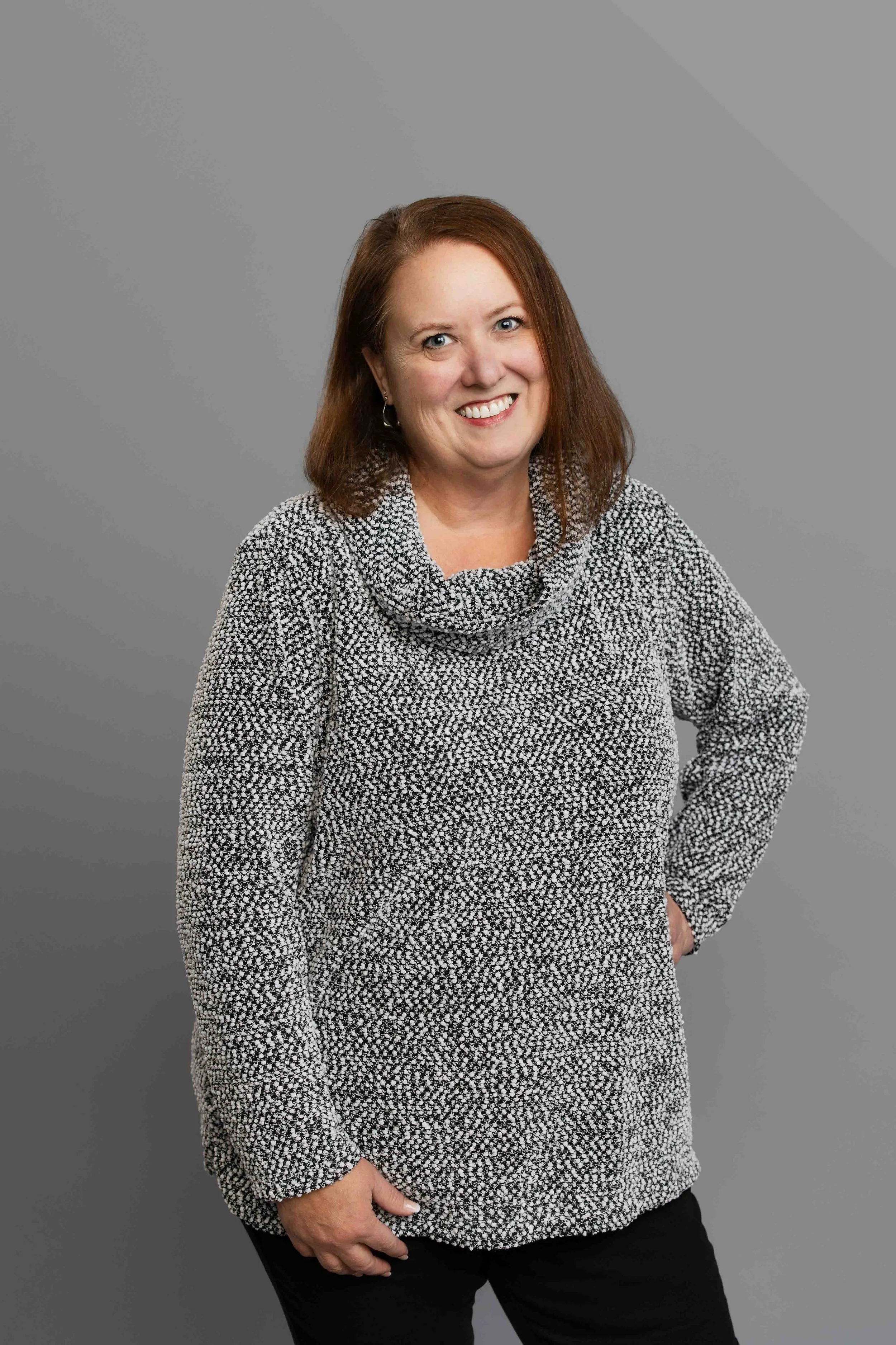 A woman with shoulder-length red hair, wearing a gray knit sweater and black pants, smiling at the camera against a plain gray background.
