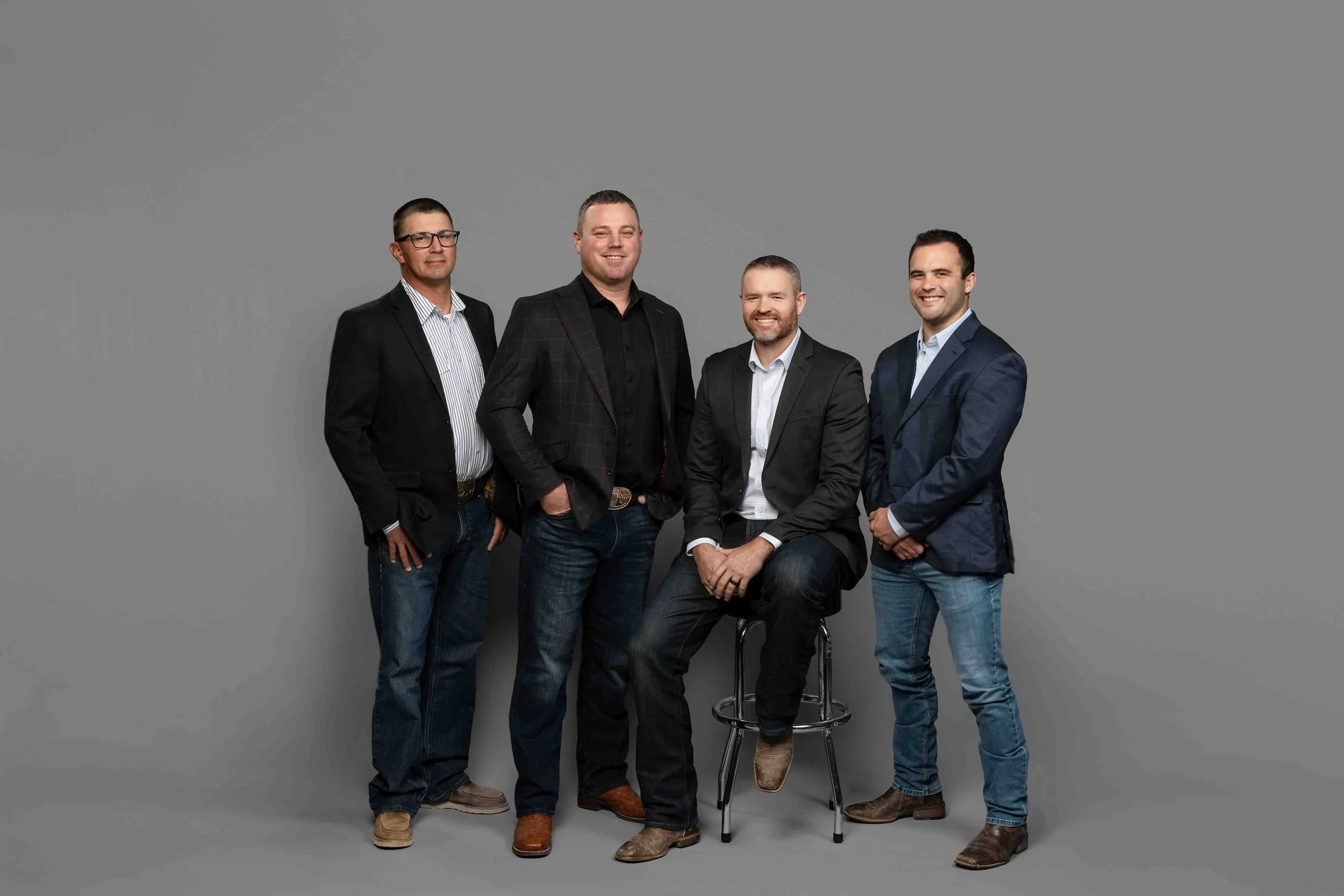 Four men in business casual attire pose together against a gray background. One is seated on a stool, and the others stand beside him, all smiling.