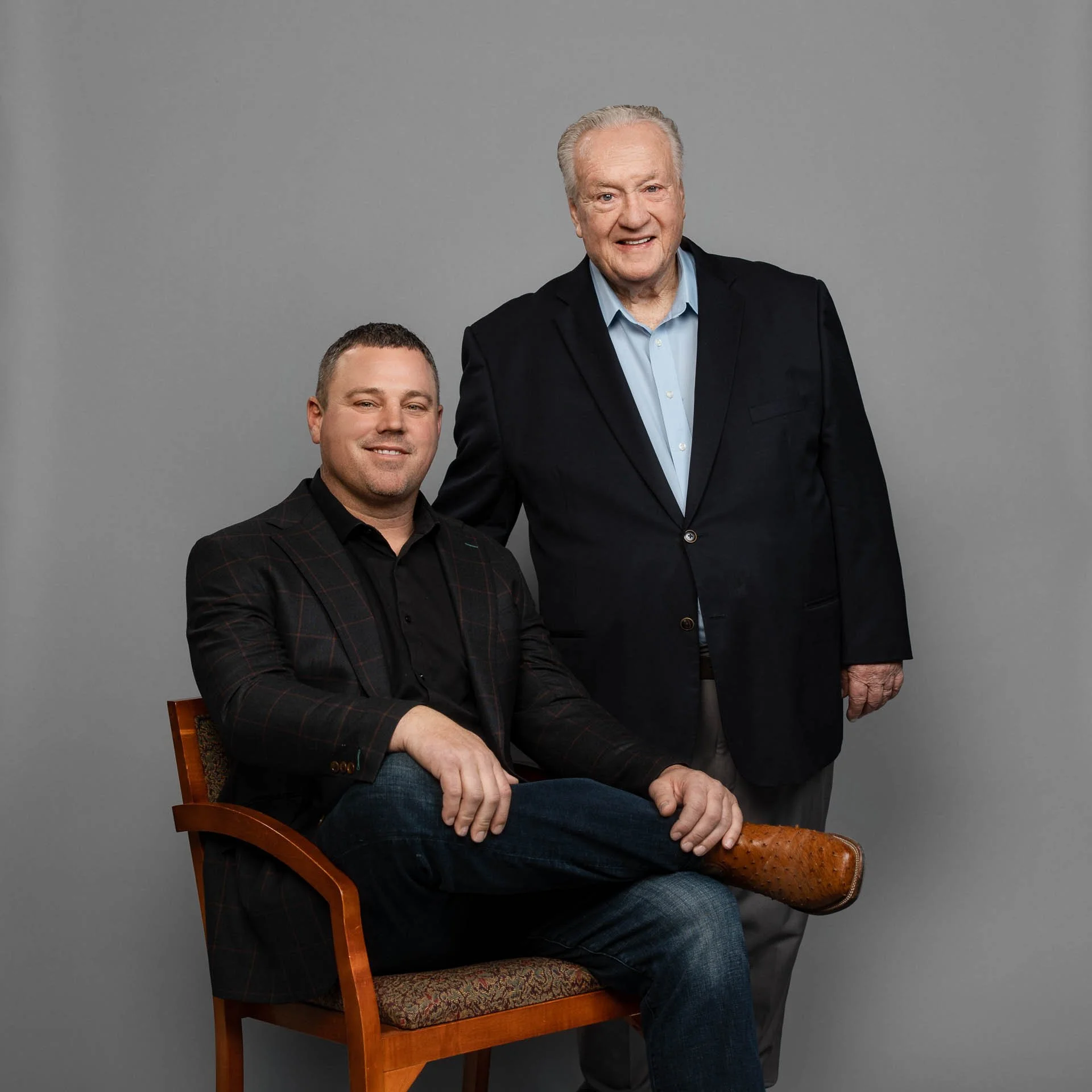 Two men posing against a gray background, one seated and one standing, both dressed in business casual attire.